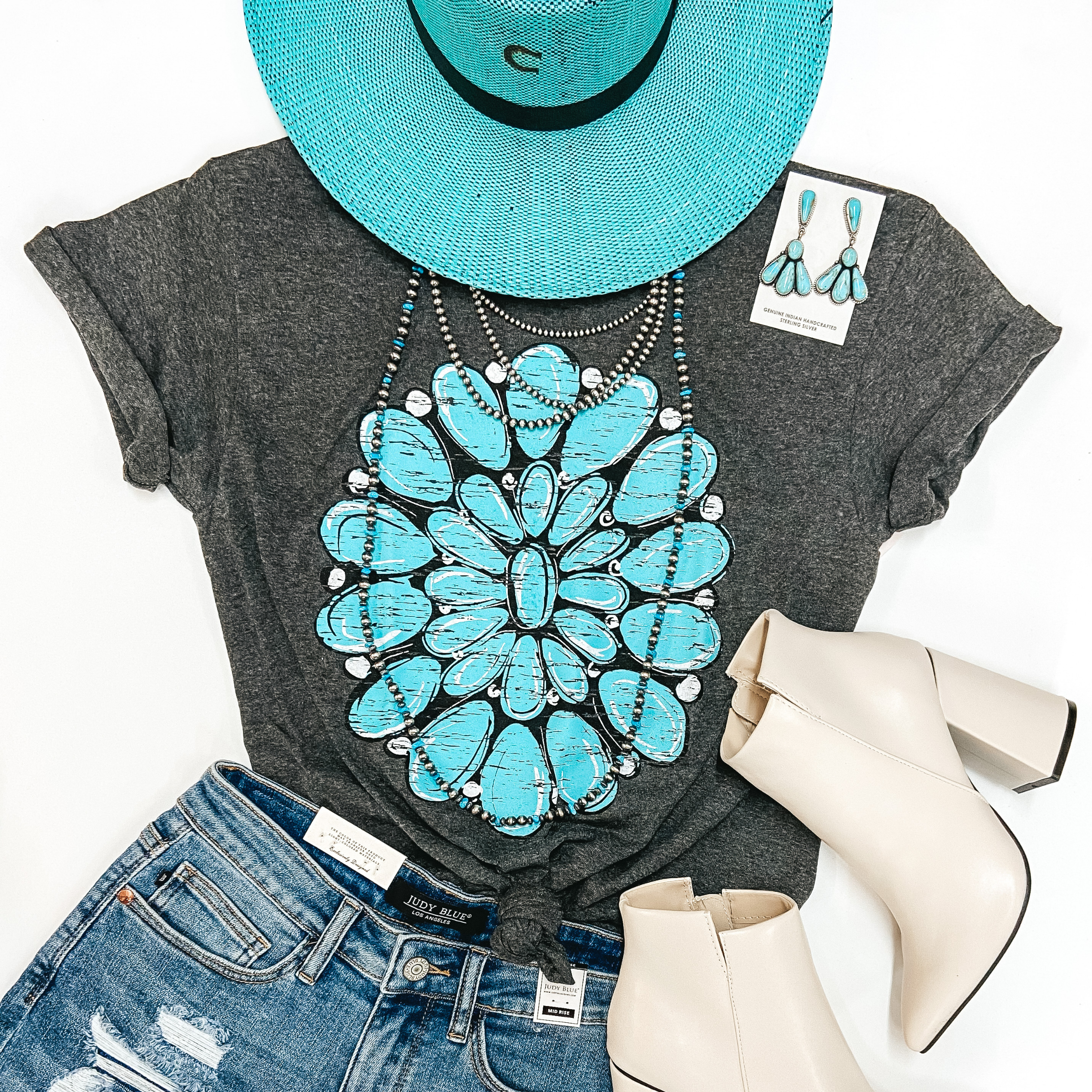 A charcoal grey tee shirt with cuffed sleeves and a knotted front. The tee shirt has a graphic of a turquoise cluster design. Tee shirt is pictured with denim shorts, silver and turquoise jewelry, ivory booties, and a turquoise straw hat.