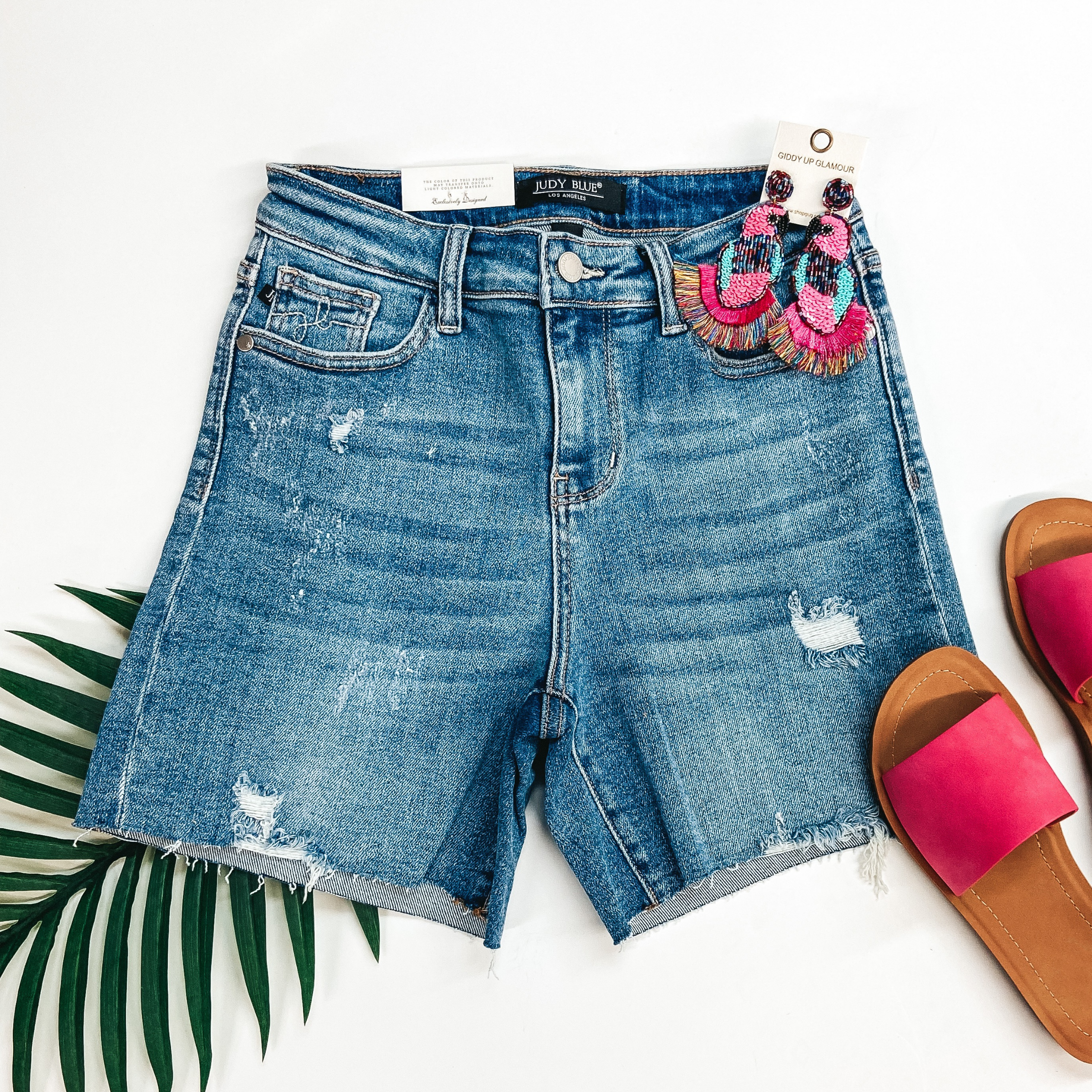 A pair of light wash distressed shorts. These shorts are pictured on a white background with a palm leaf, pink sandals, and beaded earrings.