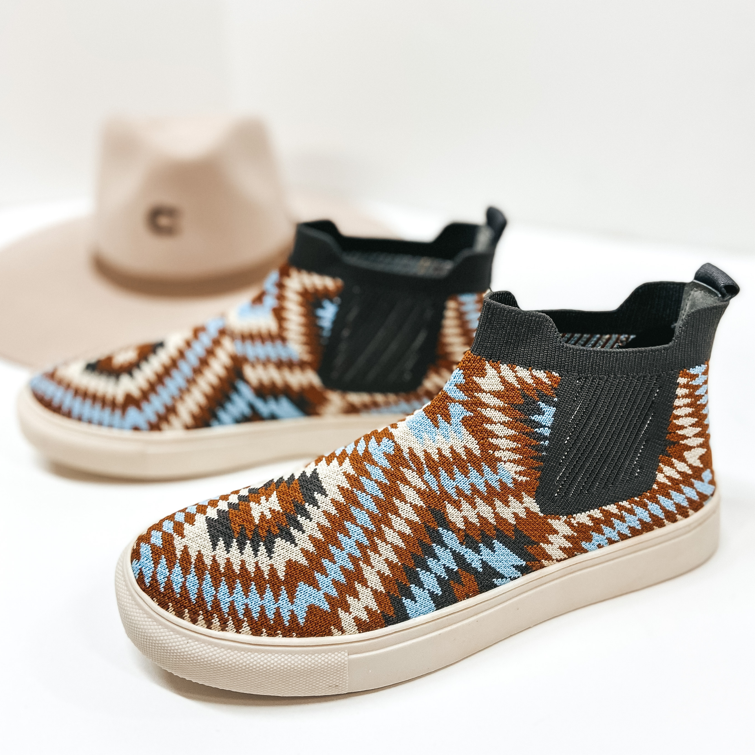Knit high top sneakers that are a tan, grey, blue, and ivory tribal print, Pictured on white background with tan hat.