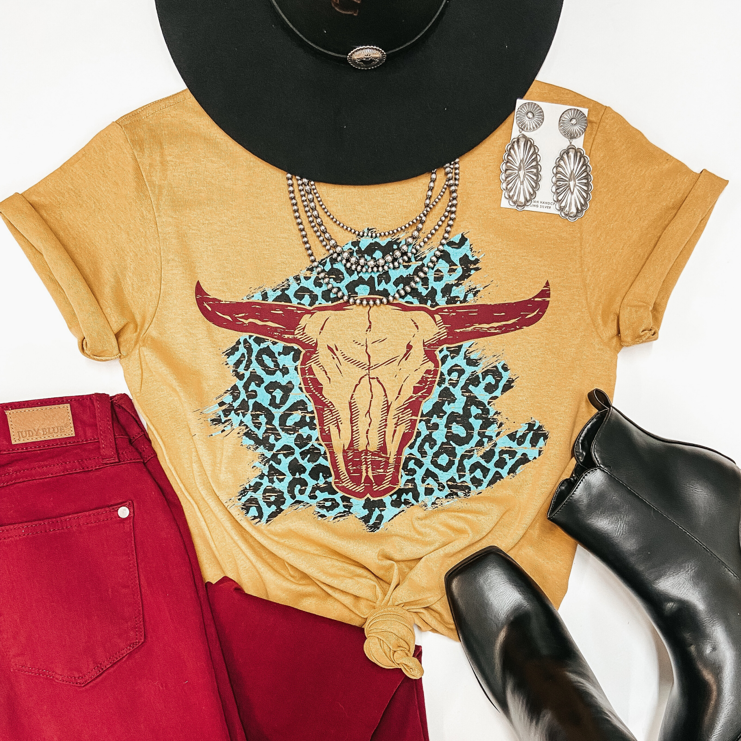 A mustard yellow graphic tee with cuffed sleeves and a knotted front. The tee shirt has a mint leopard print graphic with a bull skull on top. Pictured on white background with black hat, maroon jeans, black booties, and sterling silver jewelry.