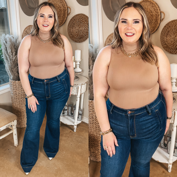 Model is wearing a turtle neck tank top that is nude. Model has it paired with skinny jeans, brown booties, and gold jewelry.