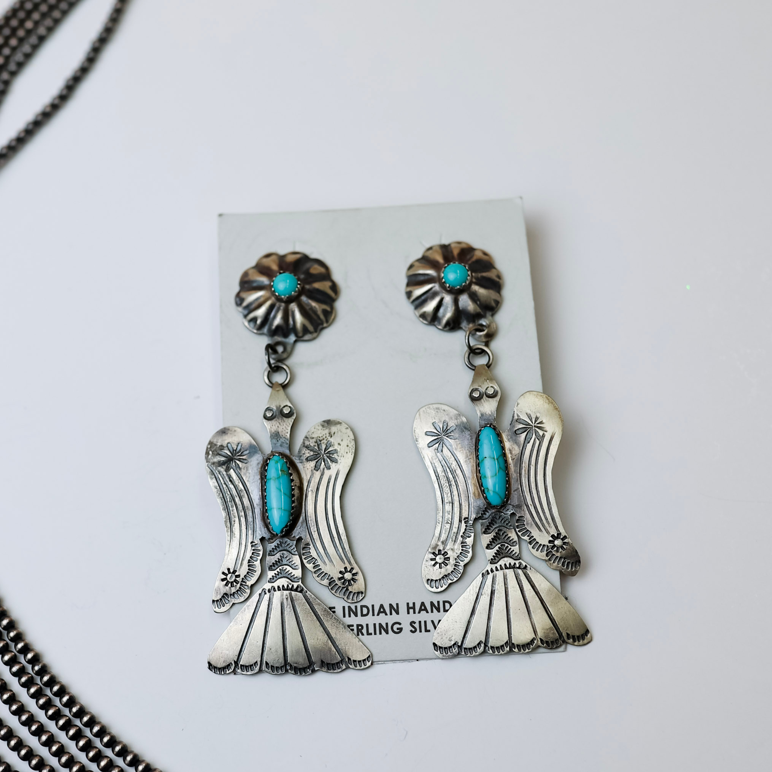 Tim Yazzie Navajo Handmade Sterling Silver with Turquoise Stones Thunderbird Dangle Earrings are centered in the middle of the picture. Navajo pearls are laid on the left side of the picture. All is on a white background. 