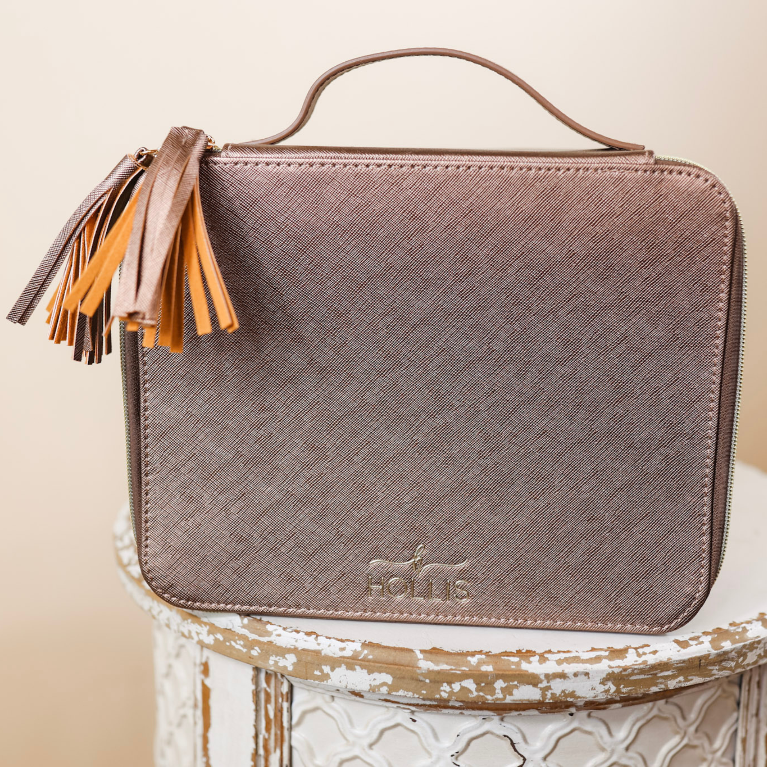 A mocha metallic bag with a handle and fringe is sitting in the center of the picture. Background is tan. 