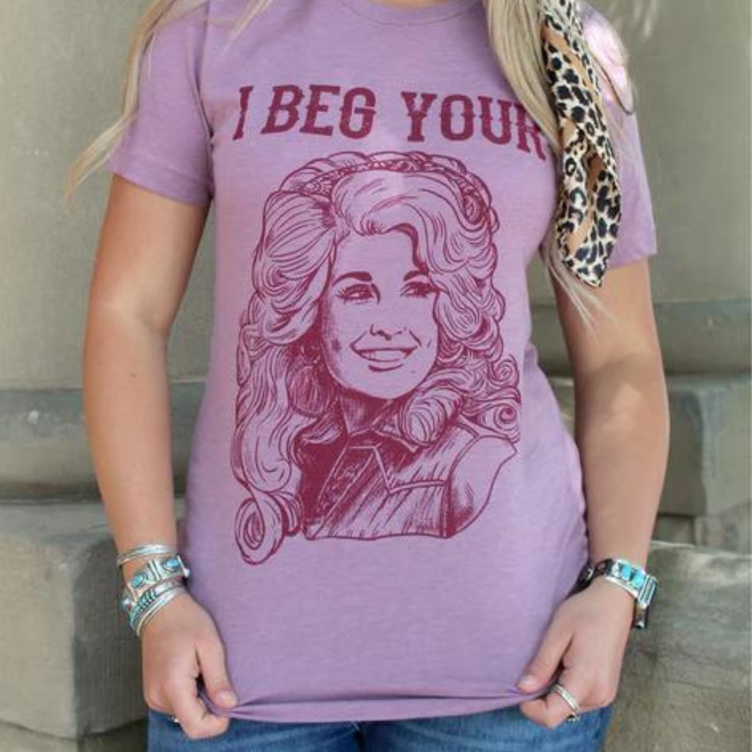 A mauve short sleeve shirt featuring a large graphic of country singer Dolly Parton in dark purple with the words "I beg your" above the image. Item is pictured on a grey background