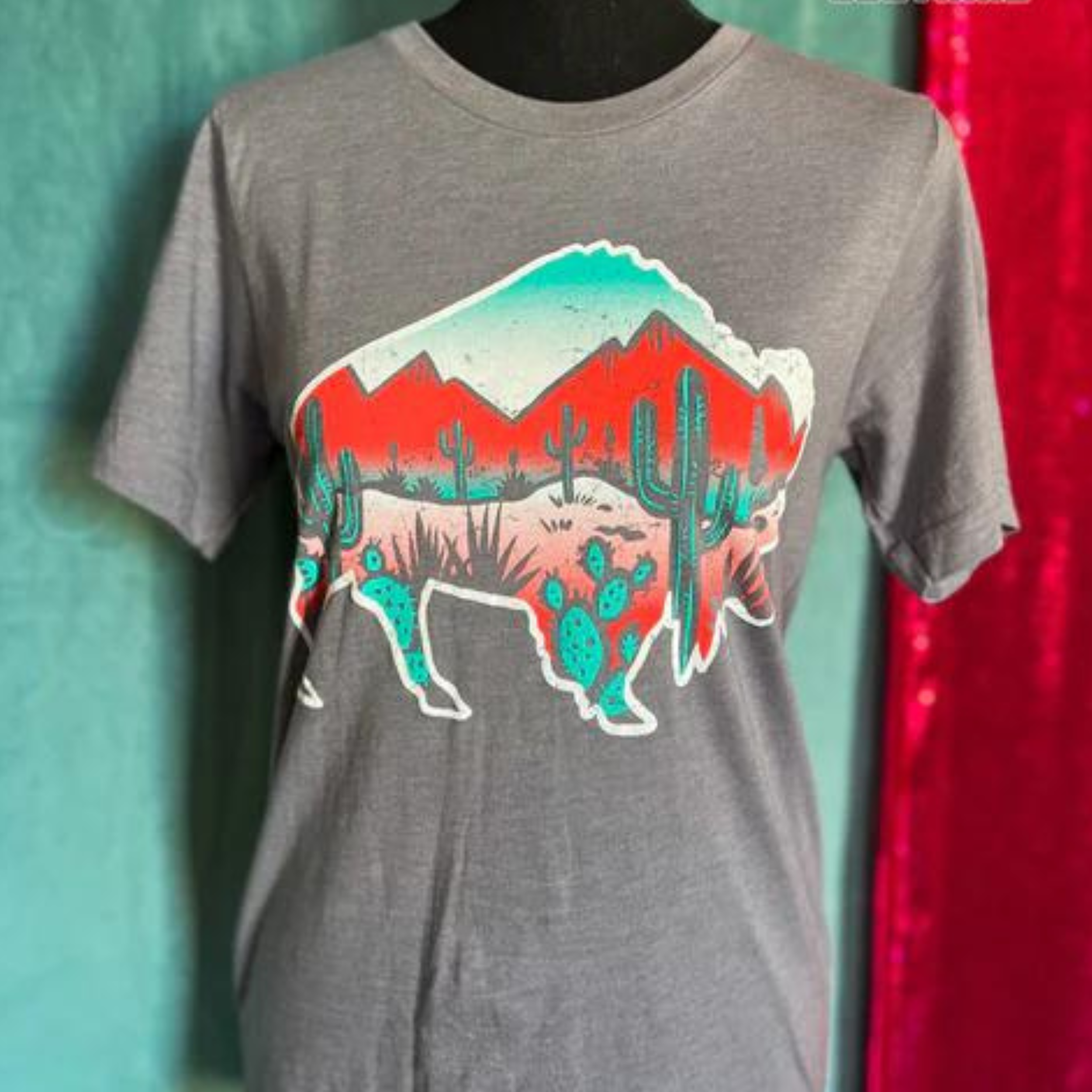 A gray short sleeve tee featuring a graphic of the outline of a bison. On the inside of the bison is a red, pink, white, and blue desert with green cacti scattered throughout and red mountains. Item is pictured on a blue and pink background.