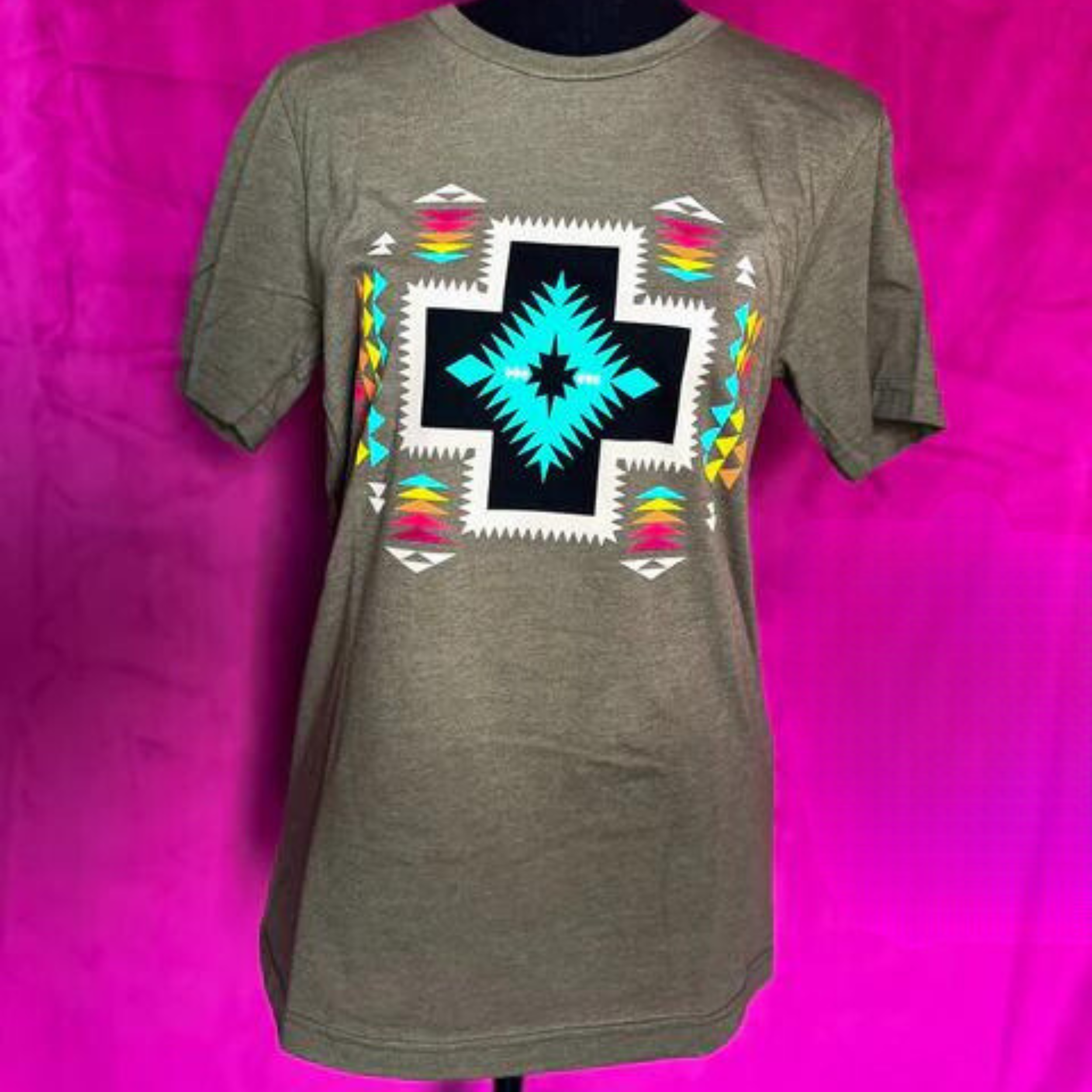 A dark heather grey tee with a tribal inspired graphic featuring a black cross with a blue angled center and angled white border. Outside of the cross is a series of rainbow arrowheads going in every direction. Item is pictured on a pink background.