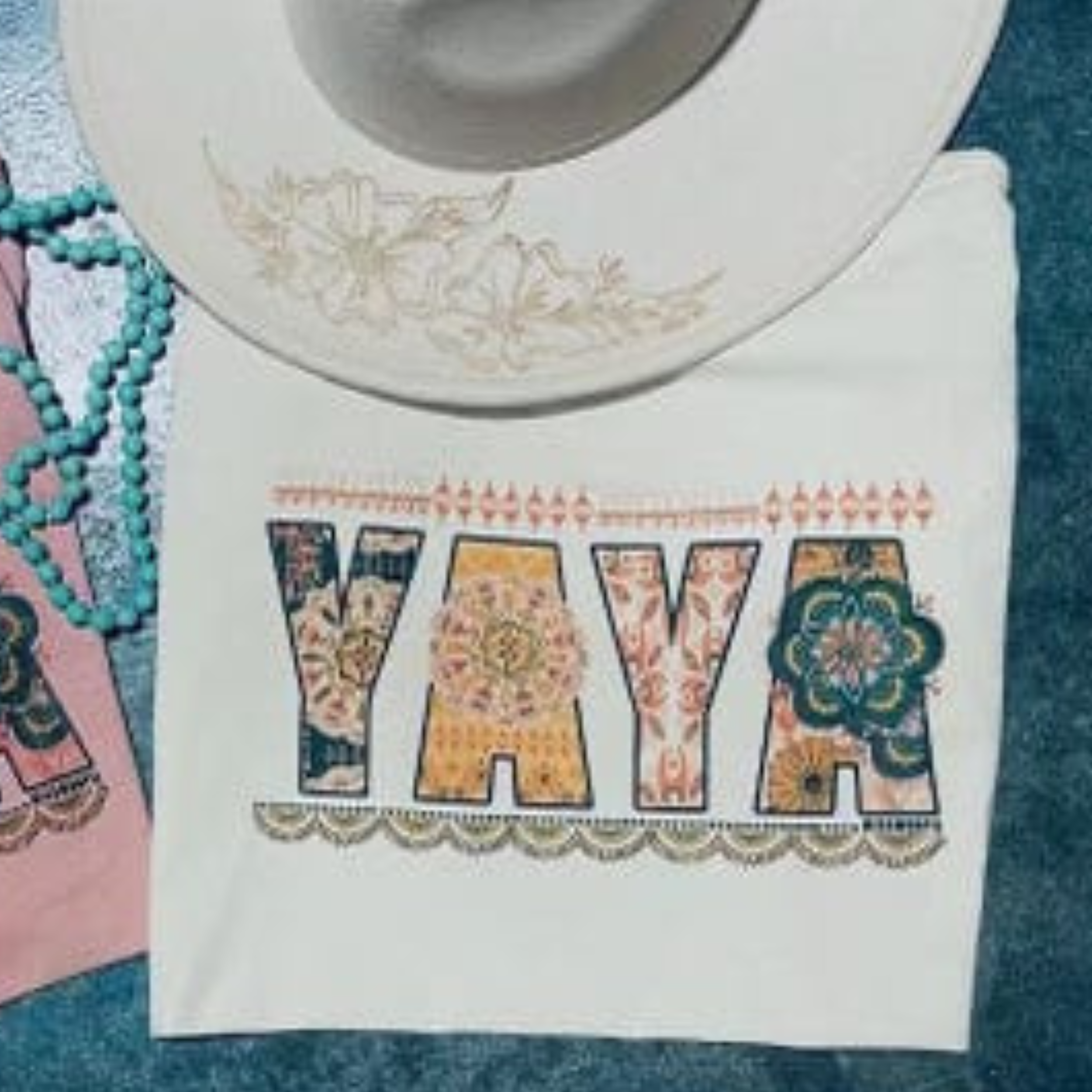 A white short sleeve tee with the word "YAYA" in the center. Each letter has its own unique pattern and color. There is a scalloped border below the text and a line border above. Item is pictured on a simple turquoise background.