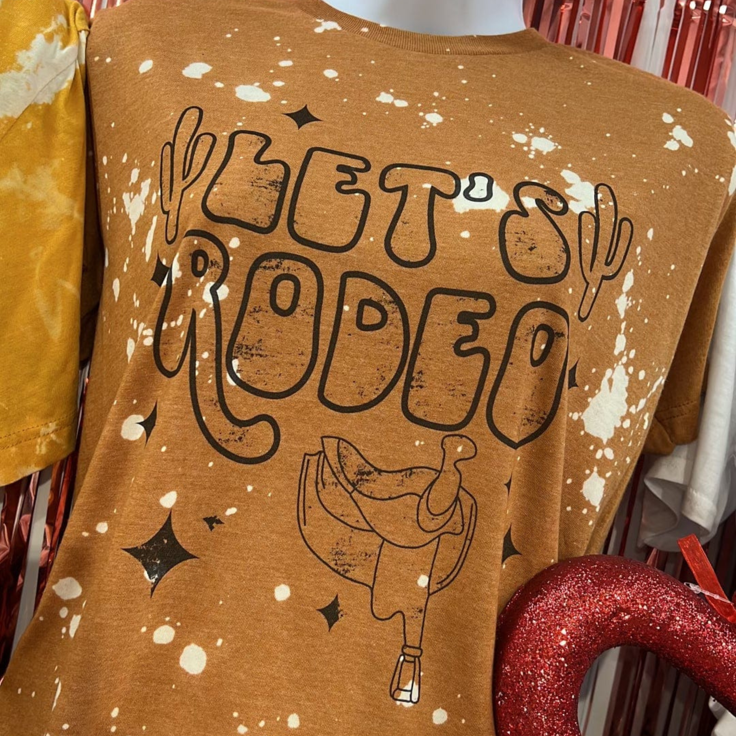 A brown short sleeve tee with a bleach splatter pattern. "Let's Rodeo" is featured in the center in outlined bubble letters. There is cacti on either side of the text, a saddle off to the right under the text, and sparkles throughout the text. Item is pictured on a multicolor background with white, yellow, and red
