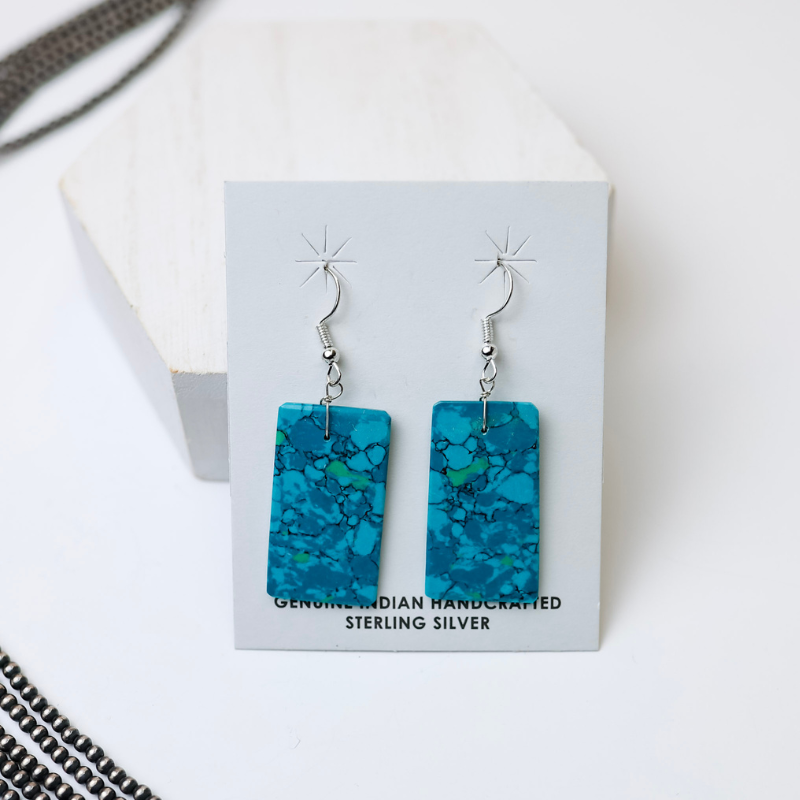 Corraine Smith Navajo Handmade Turquoise Rectangle Slab Earrings in Deep Teal are centered in the picture, with navajo pearls to the left of the earrings. All on a white background. 