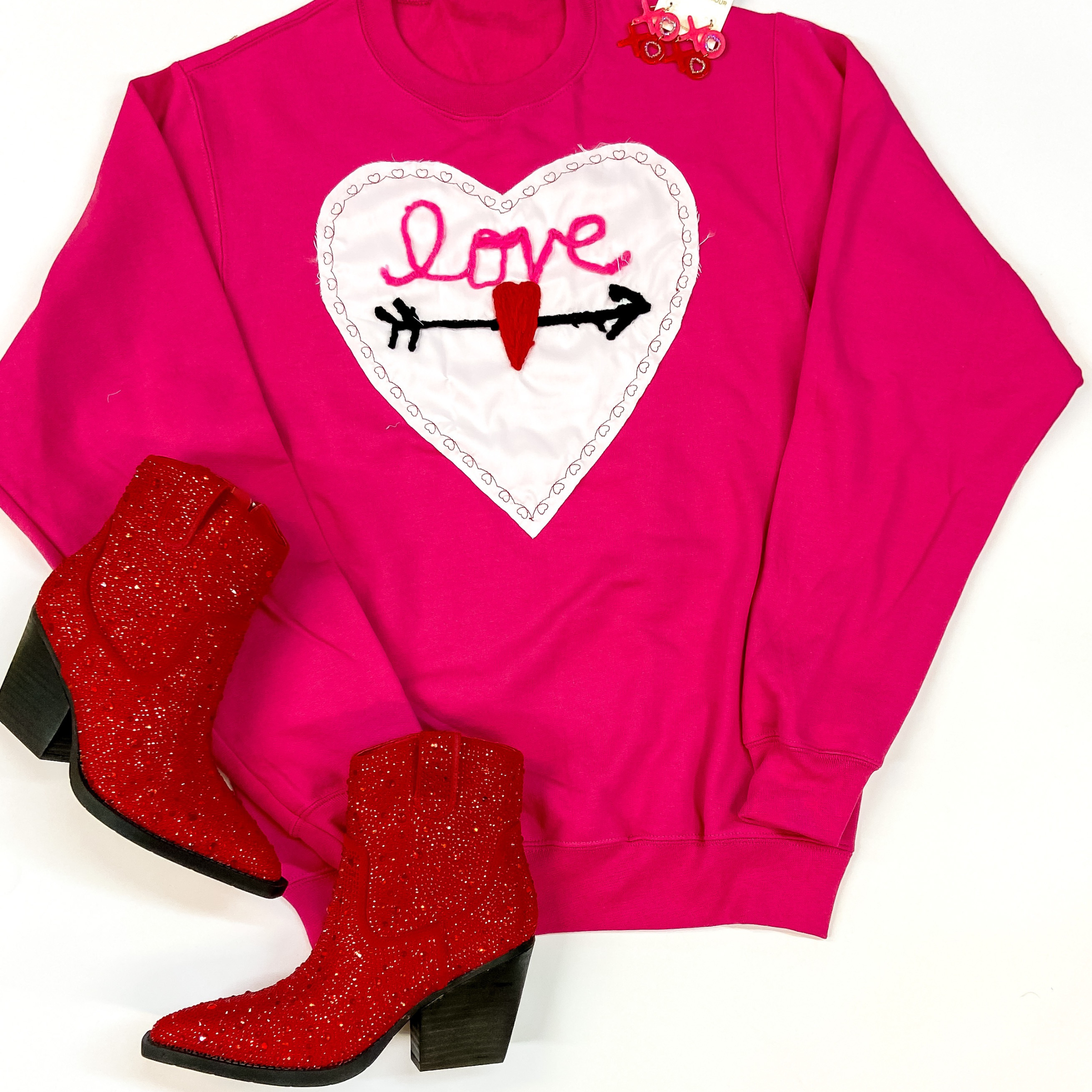 A hot pink graphic sweatshir lays in the middle of the picture with a crocheted heart in the middle of it with "LOVE" written inside the heart. Red sparkly boots lay to the left of the sweatshirt. On an all white background. 