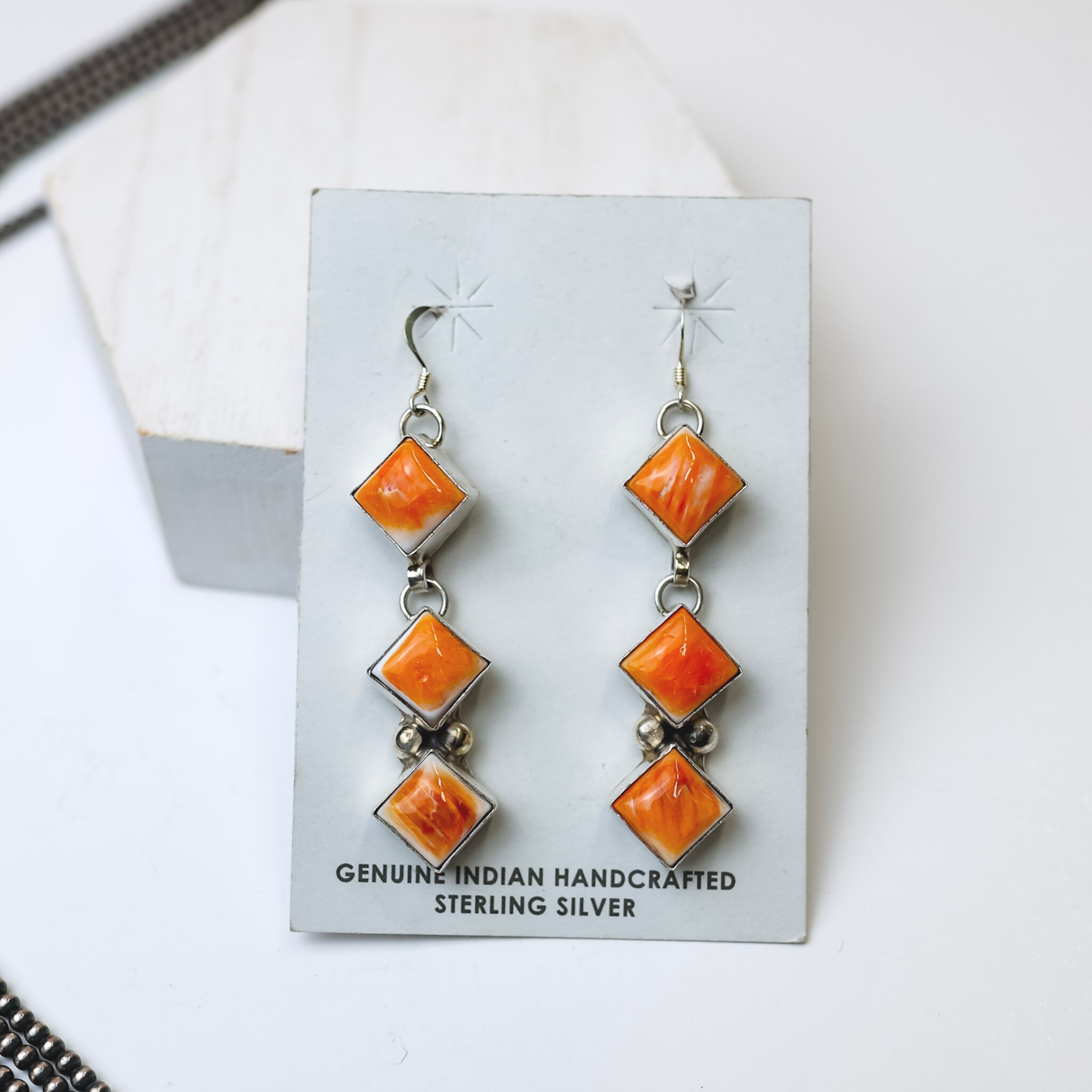 Gabby Spencer Navajo Handmade Orange Spiny Oyster Three Stone Drop Earrings are centered in the middle of the picture, with navajo pearls laid to the left of the earrings. All is on a white background. 