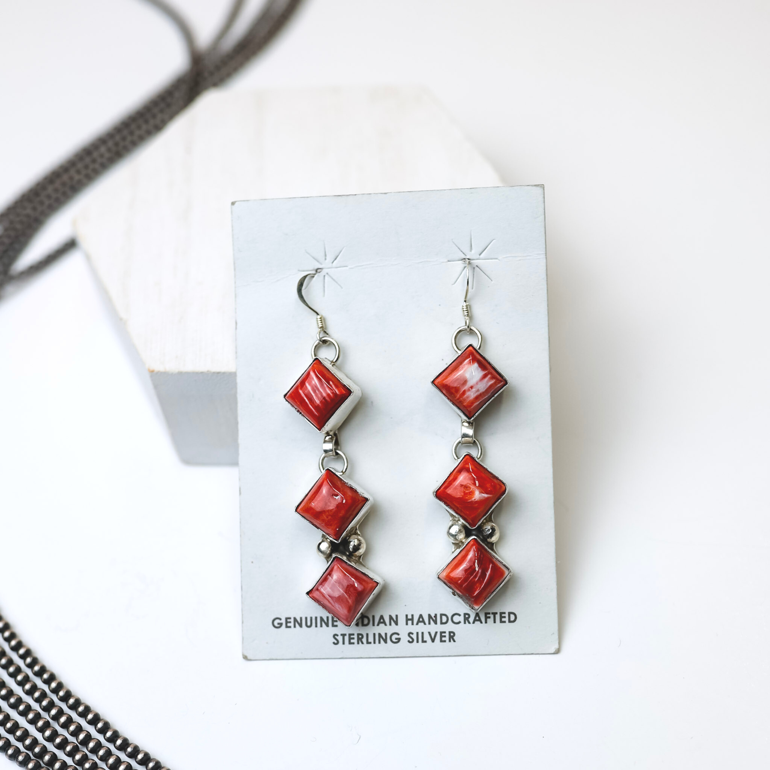 Gabby Spencer Navajo Handmade Red Spiny Oyster Three Stone Drop Earrings are centered in the picture, with navajo pearls laid on the left of the earrings. All on a white background. 