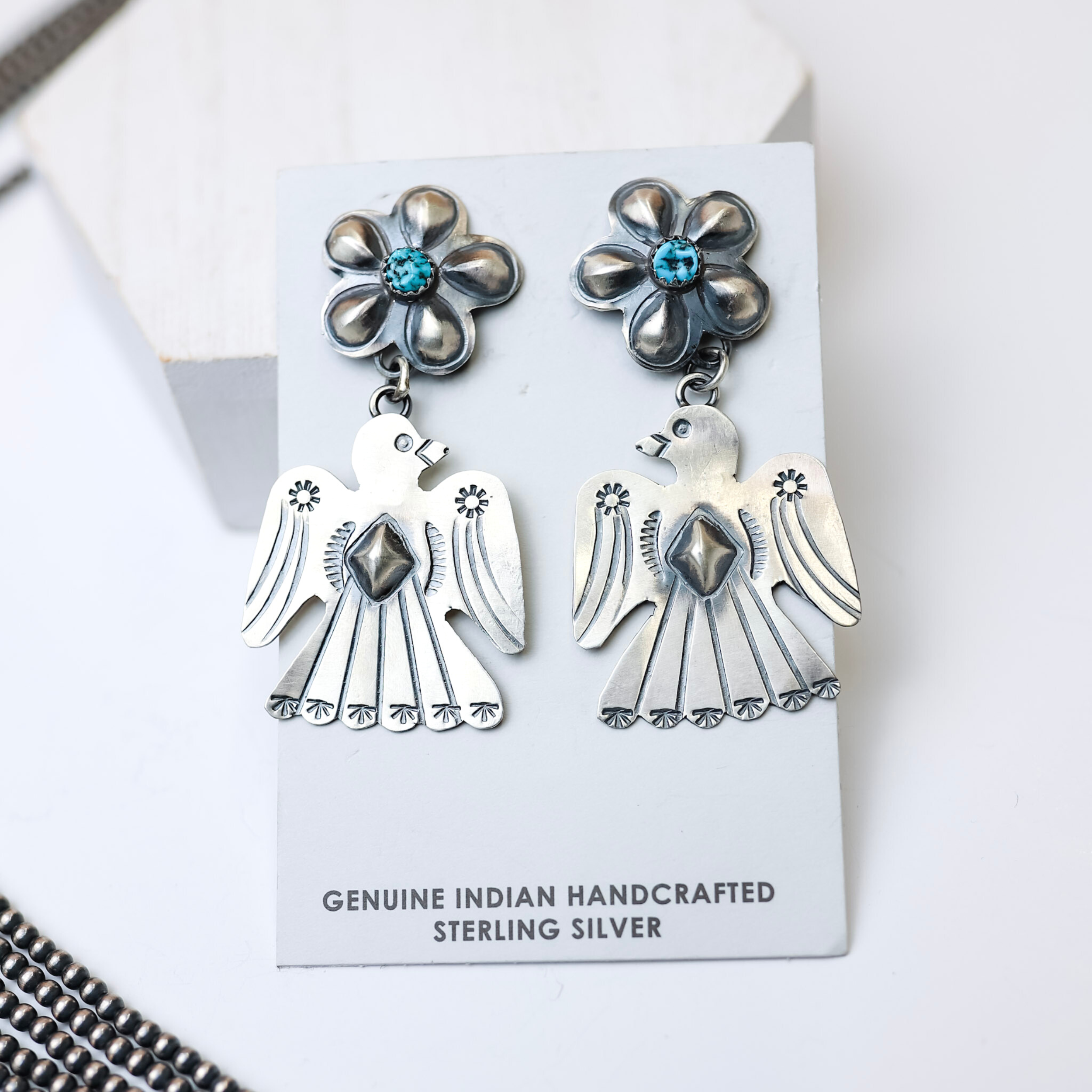Tim Yazzie Navajo Handmade Sterling Silver Flower Post Thunderbird Drop Earrings with Turquoise Stones are centered in the middle of the picture with navajo pearls to the left of the earrings. All on a white background. 