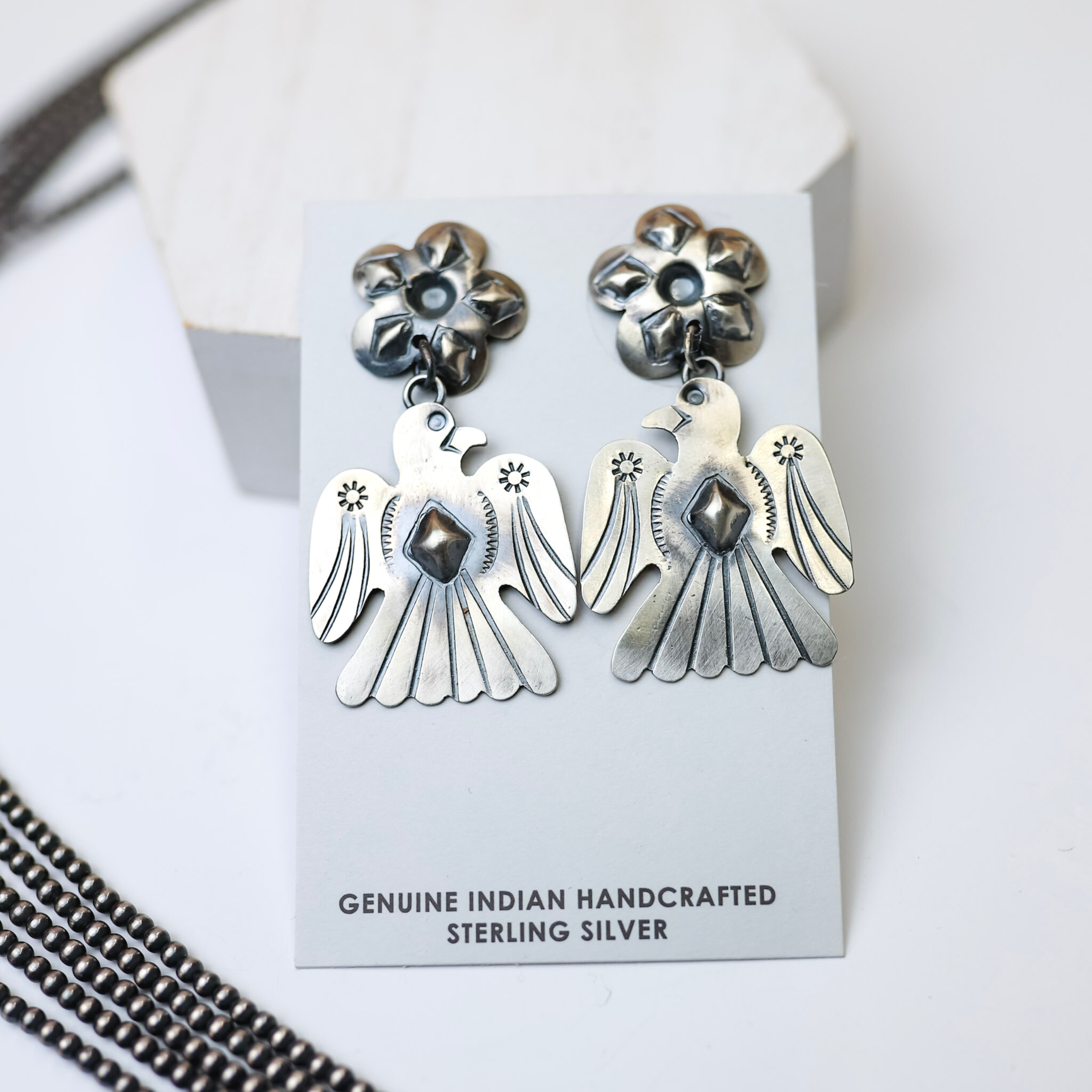 Tim Yazzie Navajo Handmade Sterling Silver Flower Post Thunderbird Drop Earrings is centered in the middle of the picture, with navajo pearls on the left of the earrings. All is on a solid white background. 