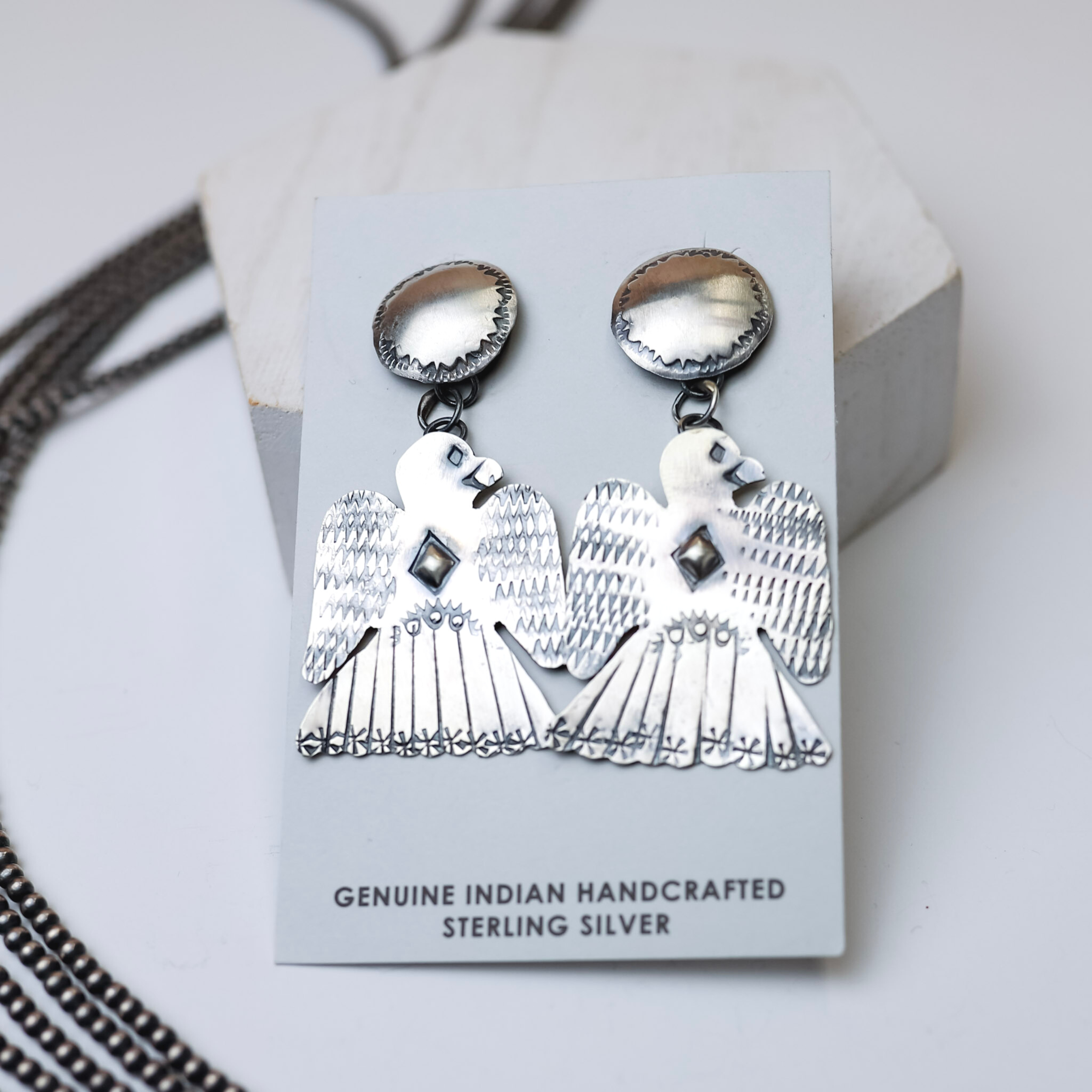 Tim Yazzie Navajo Handmade Sterling Silver Thunderbird Drop Earrings are centered in the picture with navajo pearls laid to the left of the earrings. All on a white background. 