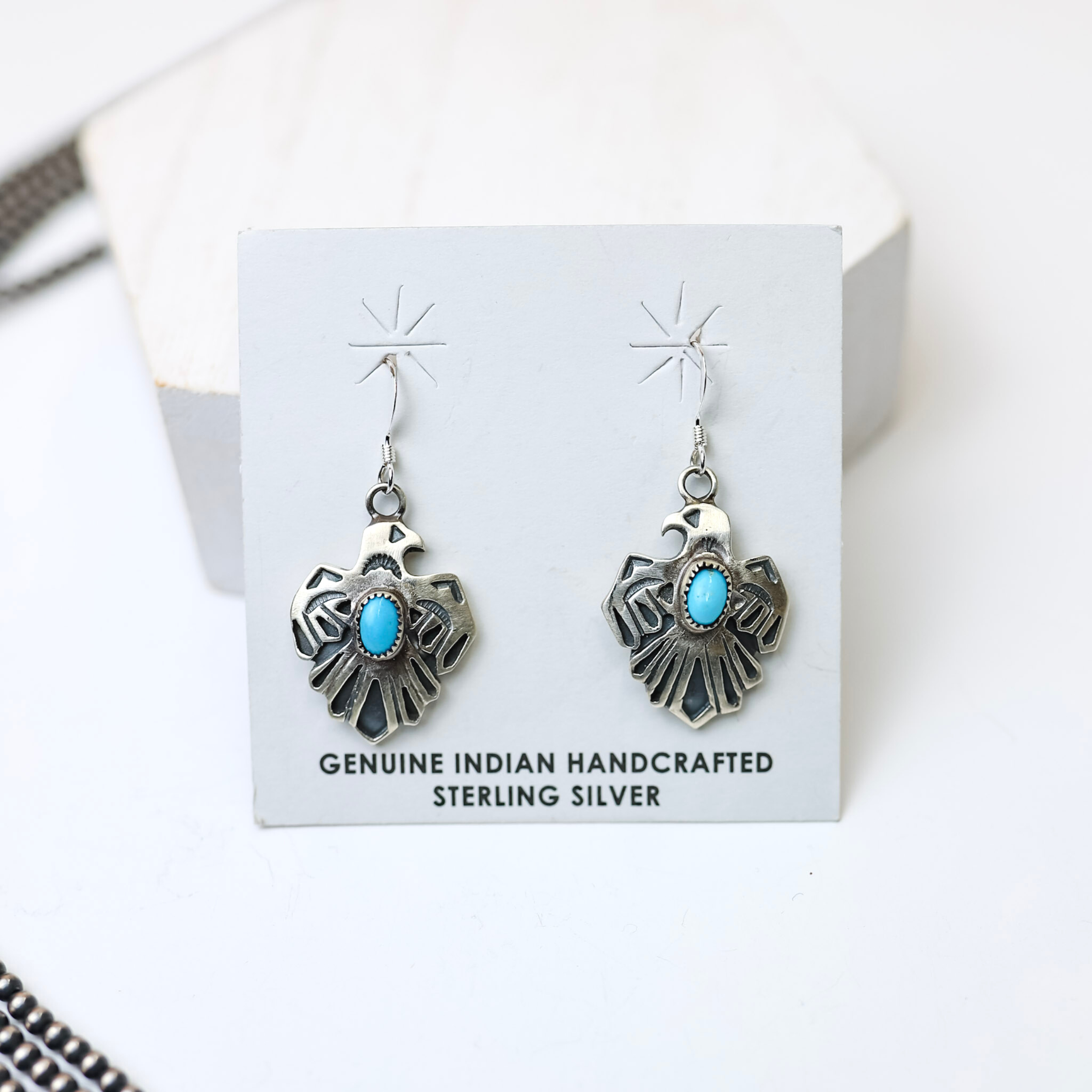 Roberta Begay Navajo Handmade Small Sterling Silver Thunderbird Earrings with Turquoise Stones is centered in the picture, with navajo pearls to the left of the earrings. All on a white background. 
