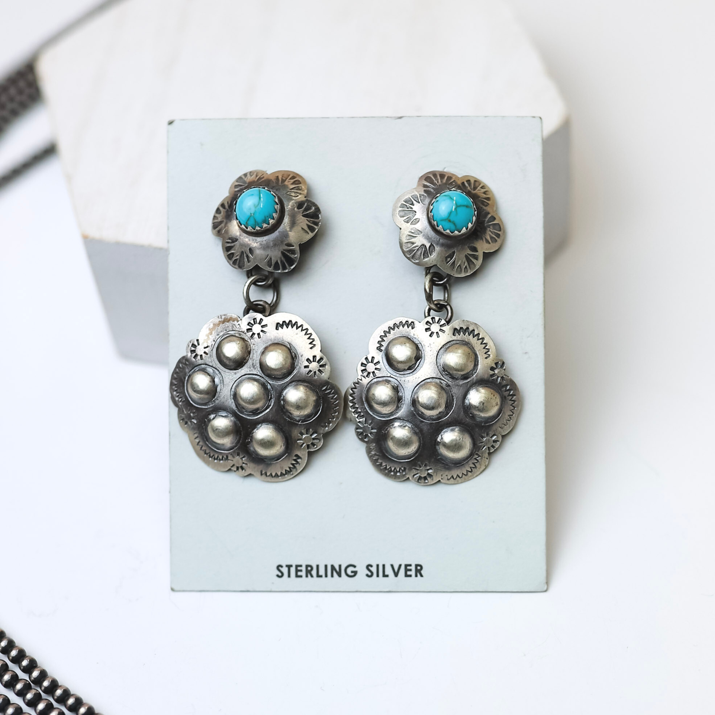 Tim Yazzie Navajo Handmade Sterling Silver Flower Drop Earrings with Turquoise Studs are centered in the picture, with navajo pearls laid to the left of the earrings. All on a white background. 