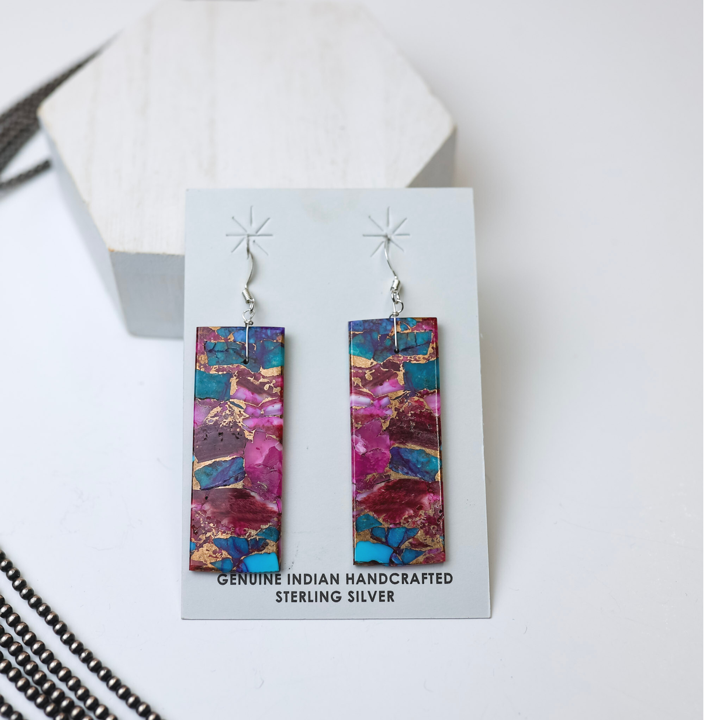 Corraine Smith Navajo Handmade Rectangle Purple Dahlia Remix Slab Earrings are centered in the picture with navajo pearls laid to the left of the earrings. All is on a white background. 