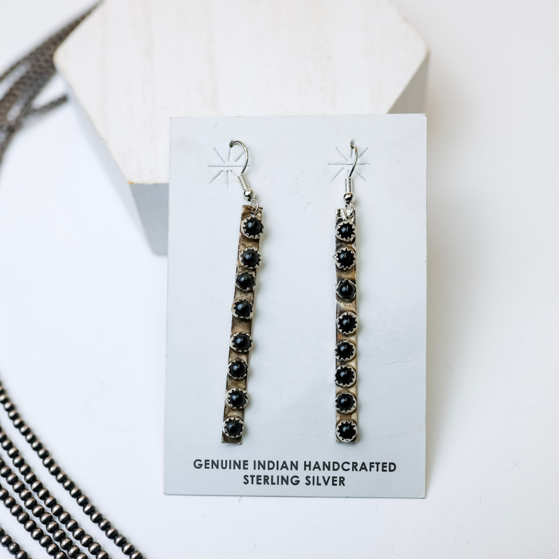 Rick Enriquez Navajo Handmade Sterling Silver Drop Earrings with Black Onyx Stones is centered in the picture, with navajo pearls laid to the left of the earrings. All is on a white background. 