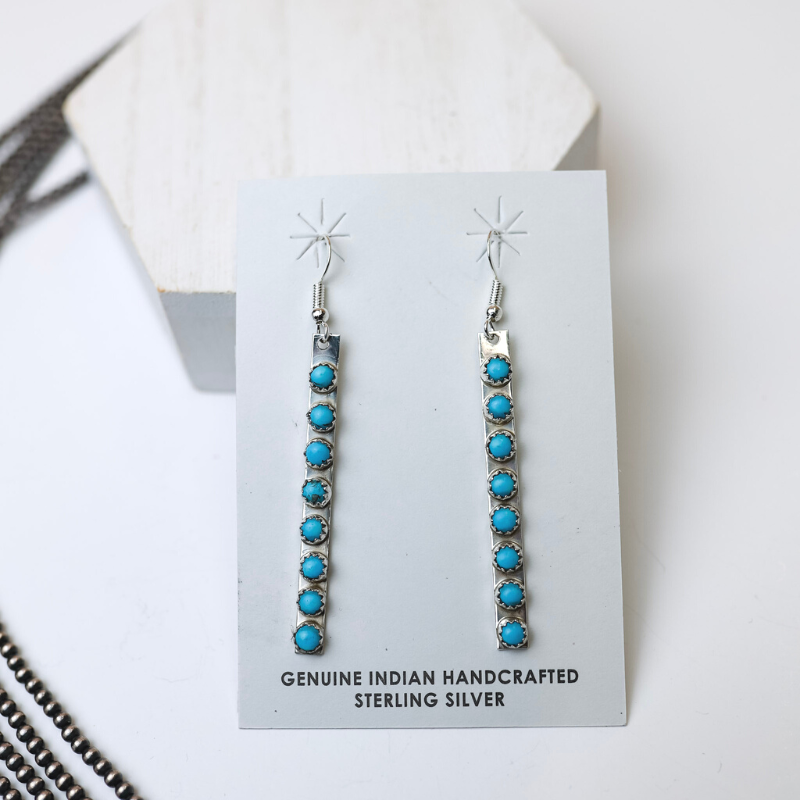 Rick Enriquez Navajo Handmade Sterling Silver Drop Earrings with Turquoise Stones are centered in the picture, with navajo pearls laid to the left of the earrings. All is on a white background. 