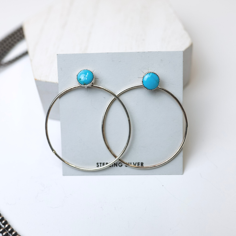 Anthony Skeets Navajo Handmade Sterling Silver Hoop Earrings with Sleeping Beauty Turquoise Studs is centered in the middle of the picture, with navajo pearls laid to the left of the earrings. All on a white background. 