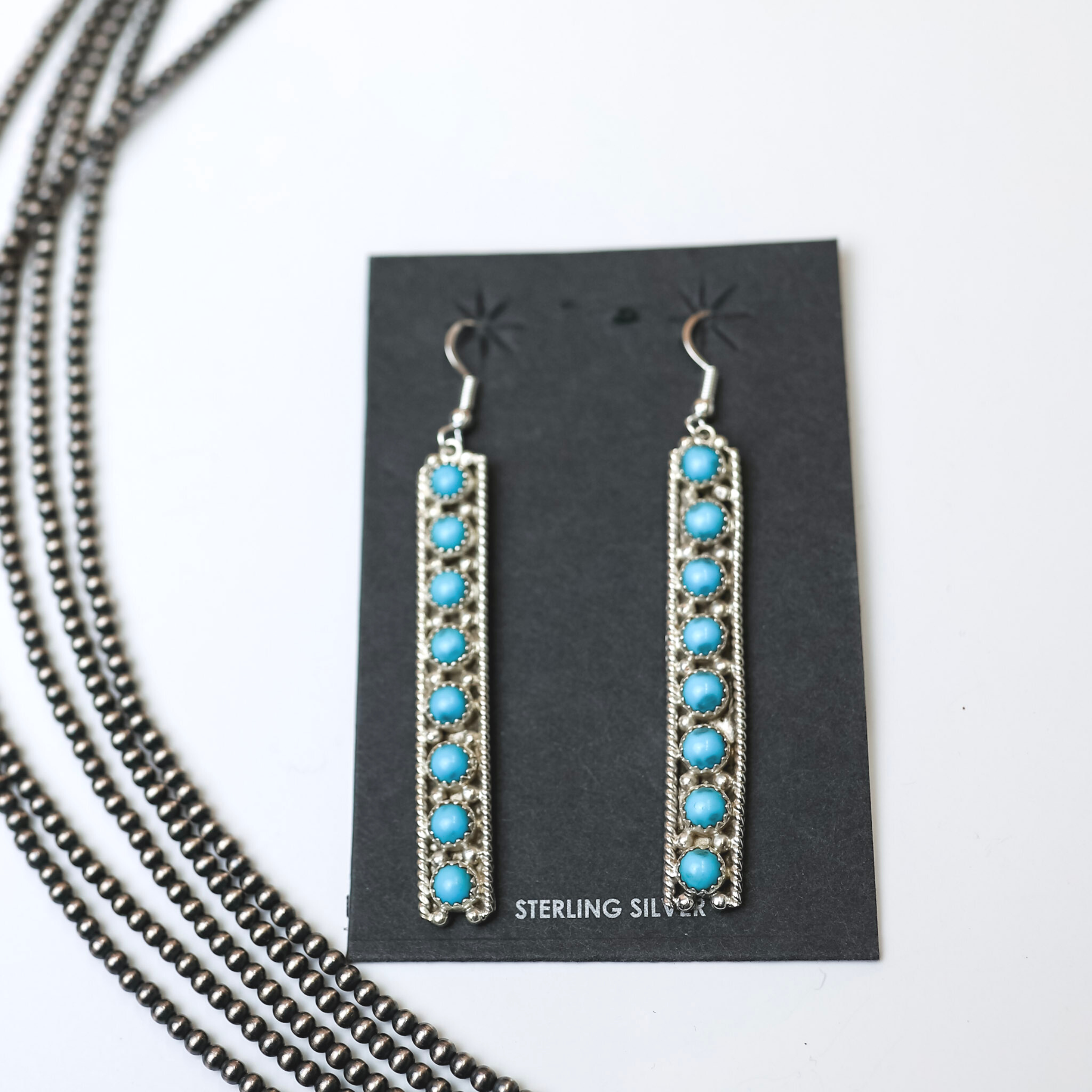 Navajo Handmade Sterling Silver Rectangle Dangle Earrings with Turquoise Stones are centered in the picture, with navajo pearls on the left side of the earrings. All the background is white.  