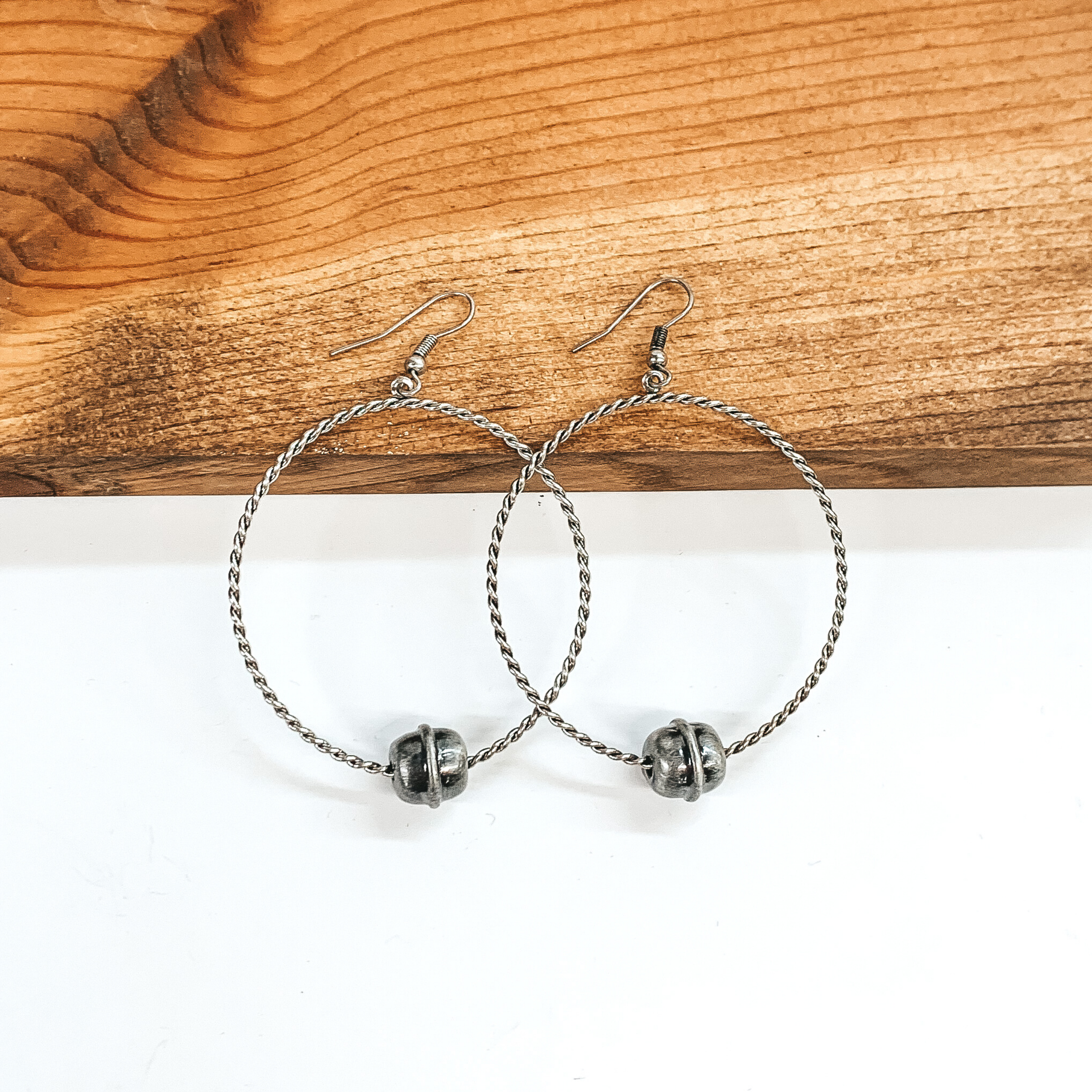 Silver twisted circle drop earrings with a single silver Navajo inspired bead at the bottom. These earrings are pictured laying against a dark piece of wood on a white background.