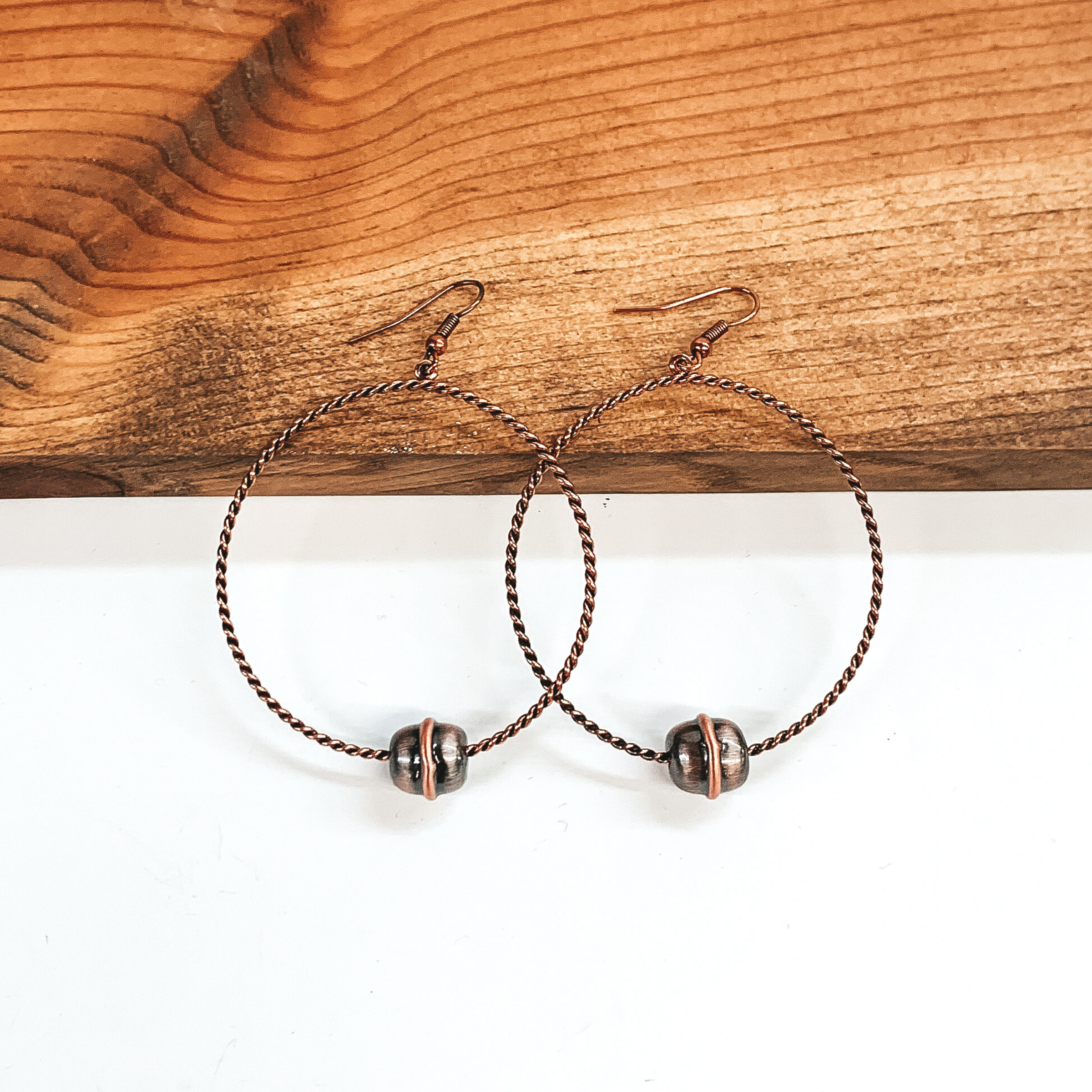Copper twisted circle drop earrings with a single copper Navajo inspired bead at the bottom. These earrings are pictured laying against a dark piece of wood on a white background.