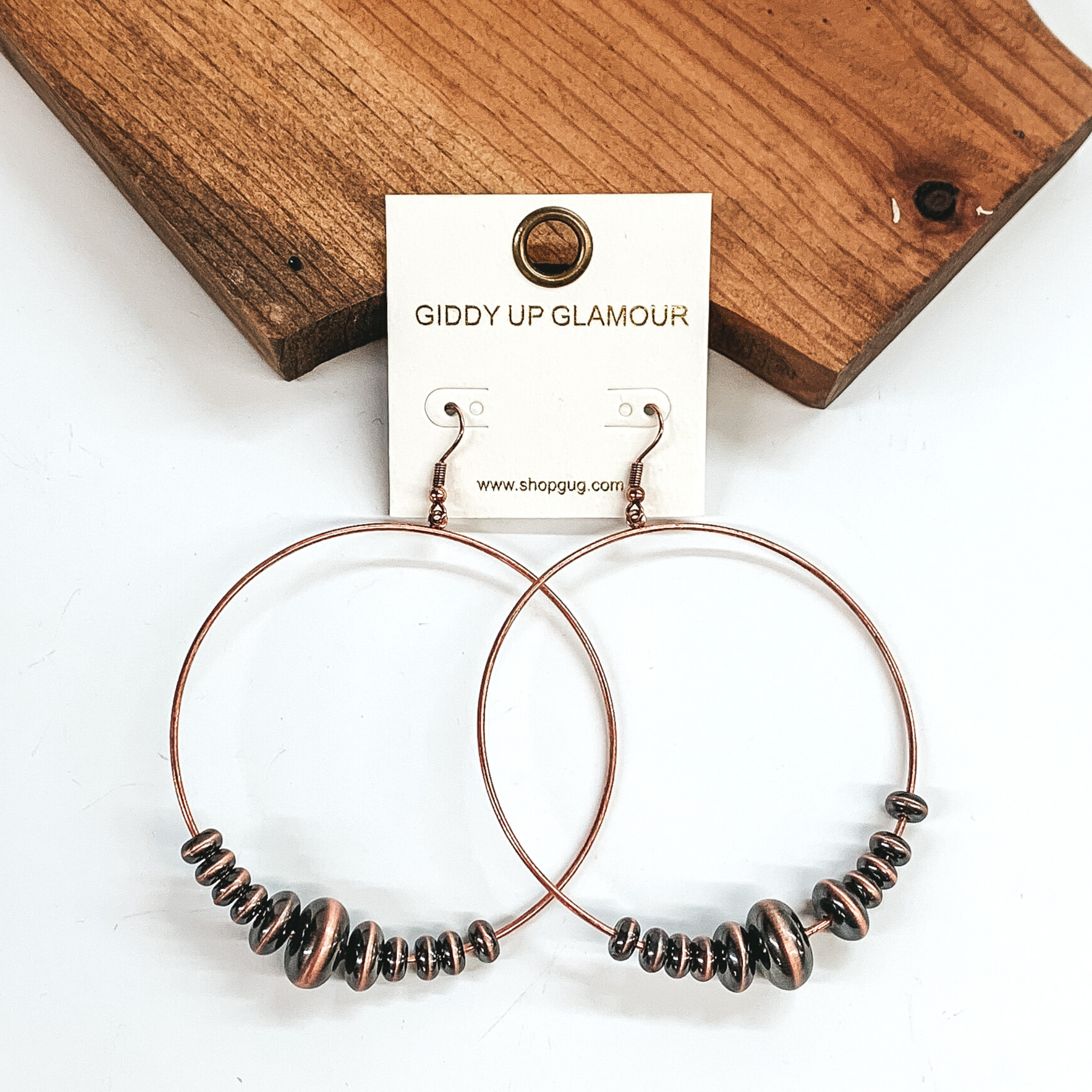 Copper open circle drop earrings with copper saucer beads in varying sizes at the bottom of the earrings. These earrings are laying against a dark piece of wood on a white background. 