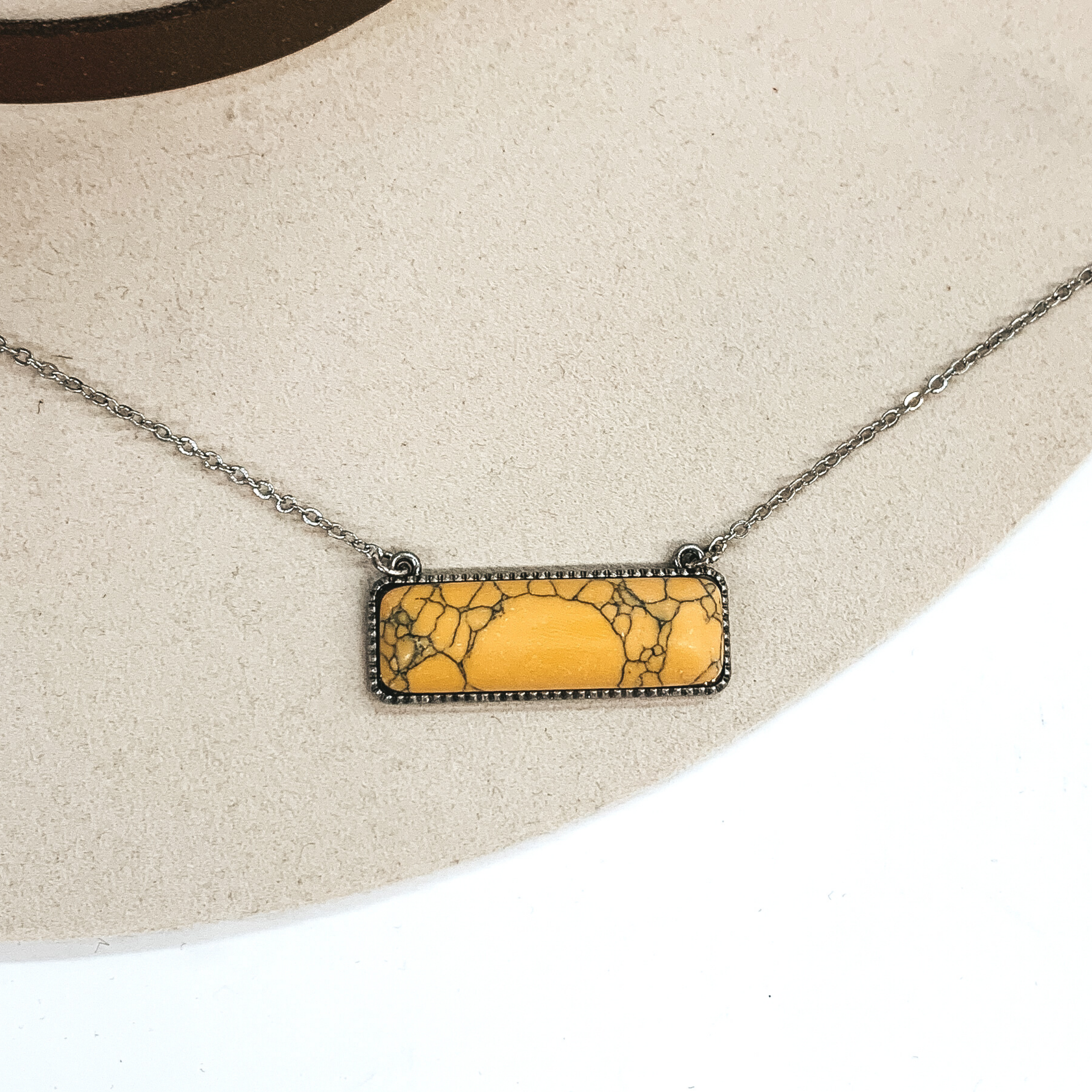 Silver chained necklace with a rectangle bar pendant. The pendant includes a yellow rectangle stone. This necklace is pictured on a white and ivory background.