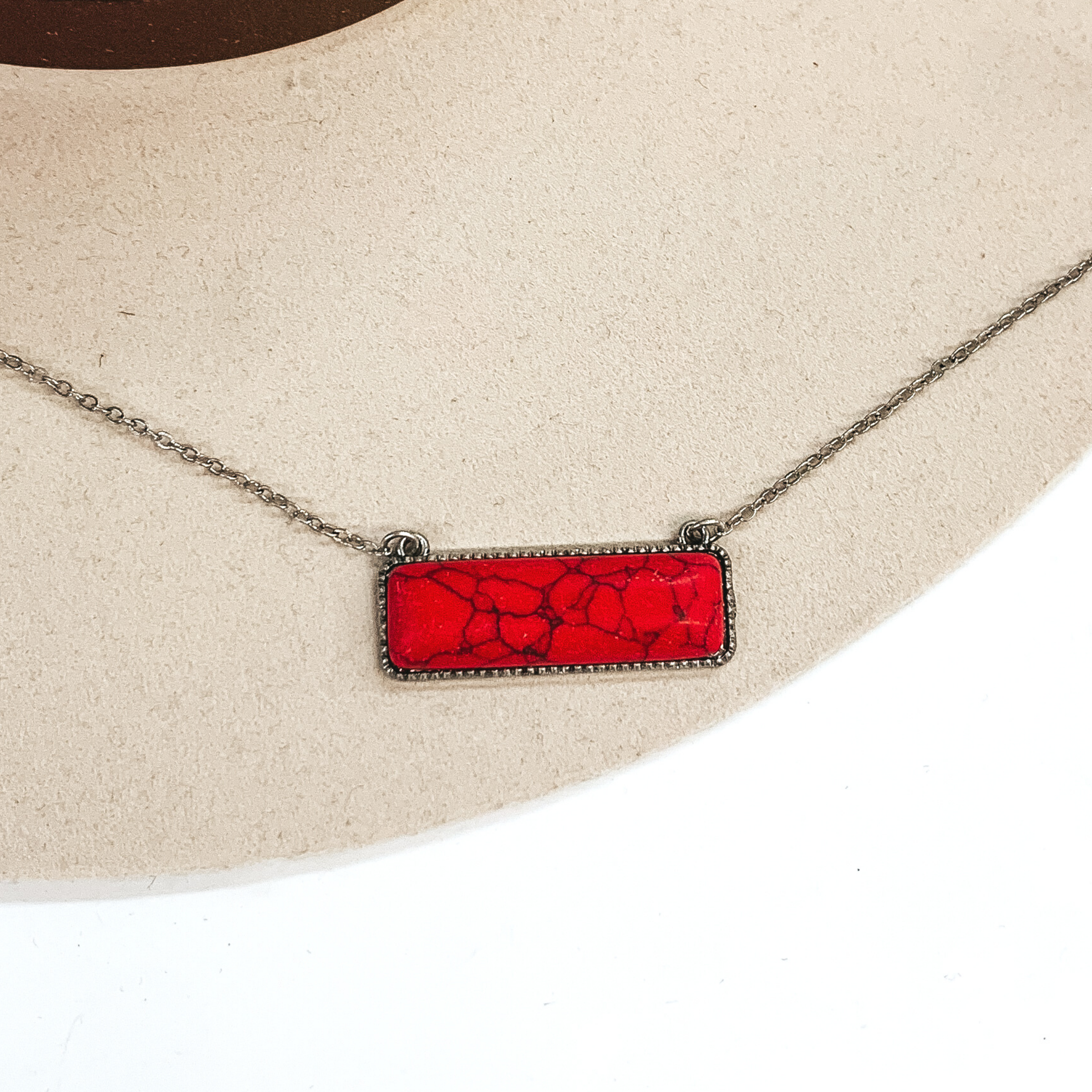 Silver chained necklace with a rectangle bar pendant. The pendant includes a red rectangle stone. This necklace is pictured on a white and ivory background.