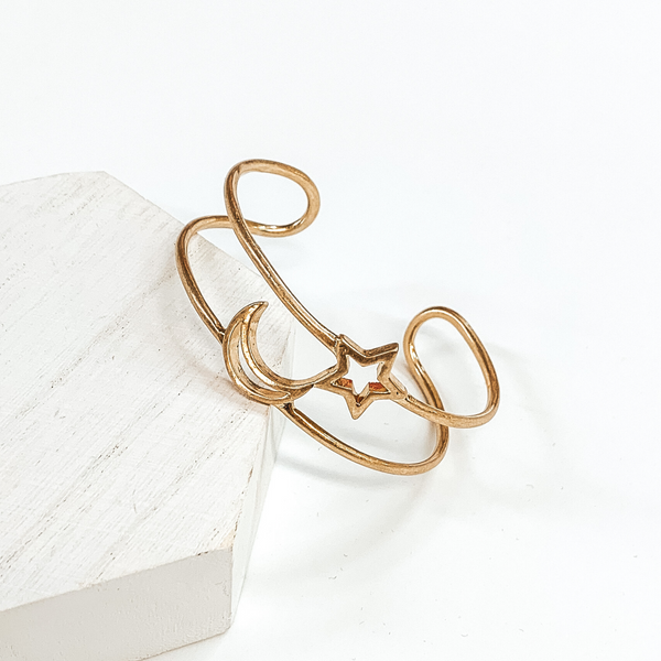 Gold outlined bangle with a moon and star outline pendant in the center of the bangle. This bracelet is pictured on a white background laying against a white piece of wood. 