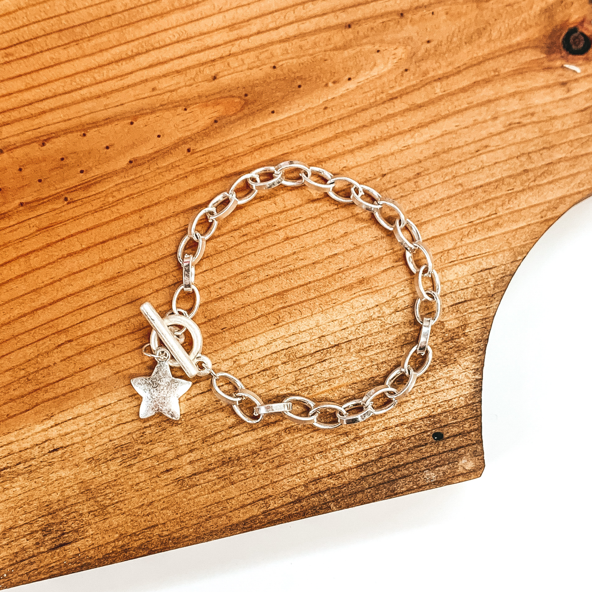 Silver oval chain linked bracelet with a toggle clasp and hanging silver star charm.  this bracelet is pictured laying on a dark brown piece of wood on a white background. 