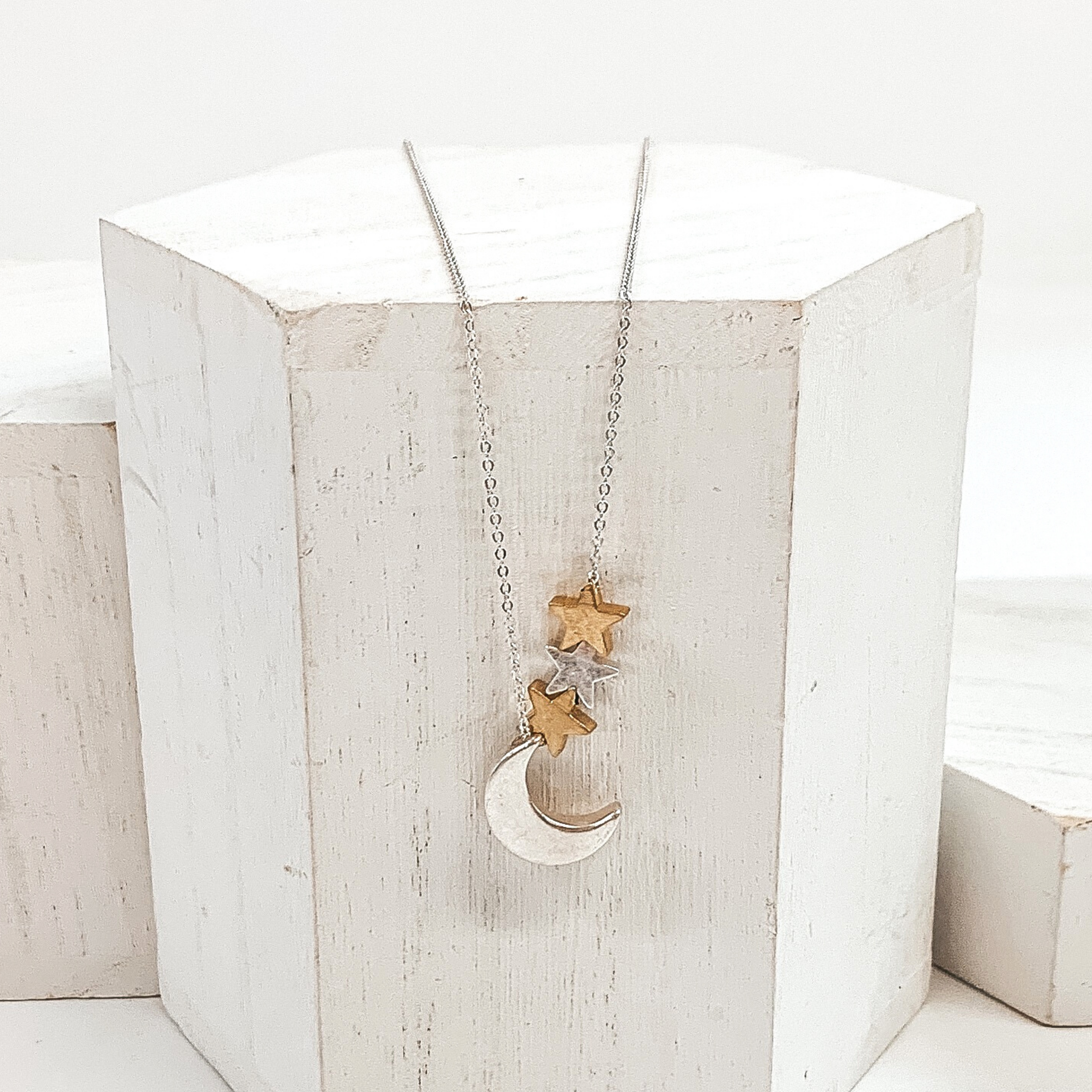 Simple silver necklace with silver moon charm, one silver star charm, and two gold star charms. This necklace is pictured on a white block on a white background.