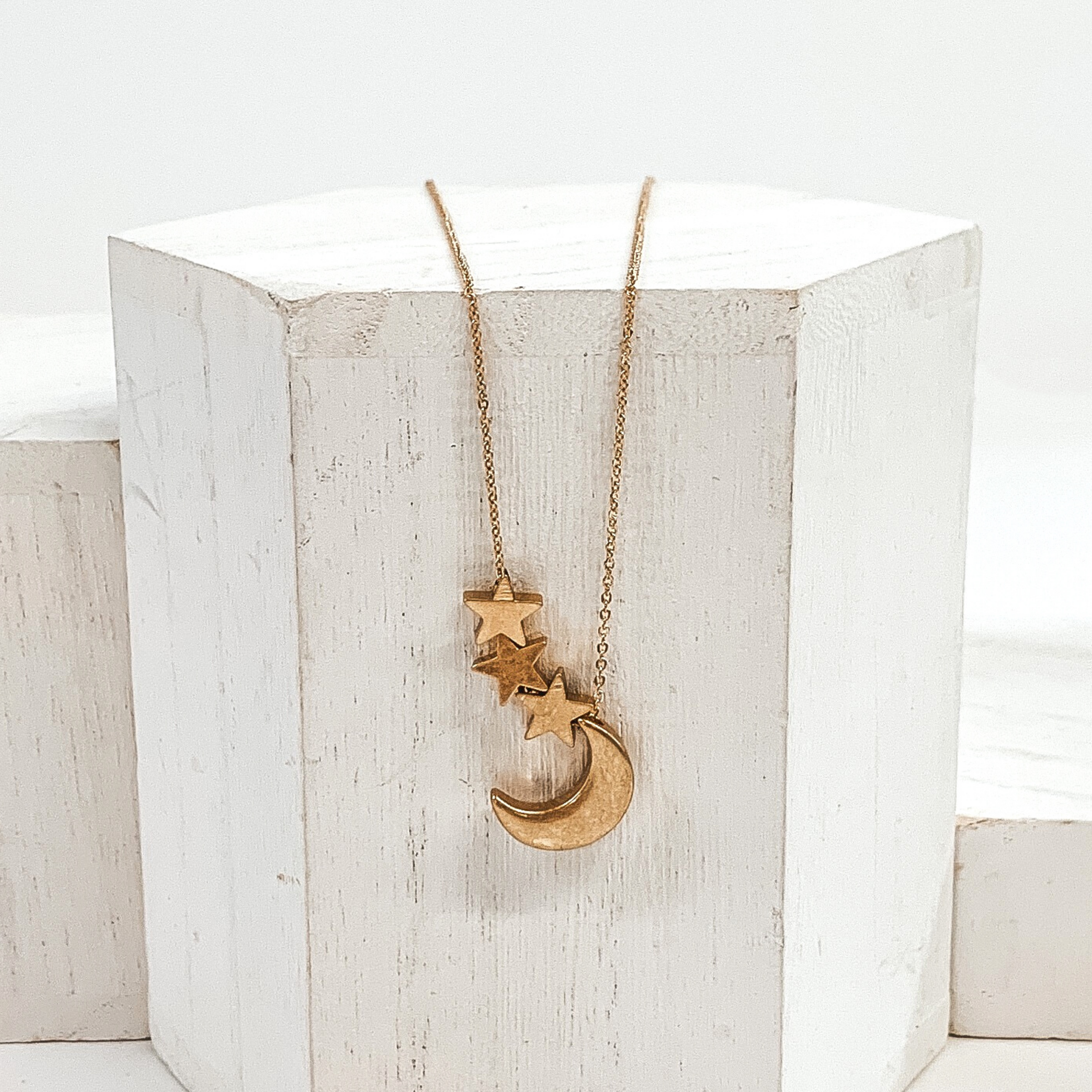 Simple gold necklace with gold moon charm and three gold star charms. This necklace is pictured on a white block on a white background.