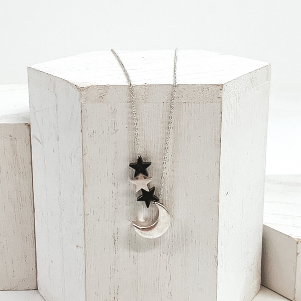 Simple silver necklace with silver moon charm, one silver star charm, and two gunmetal star charms. This necklace is pictured on a white block on a white background. 