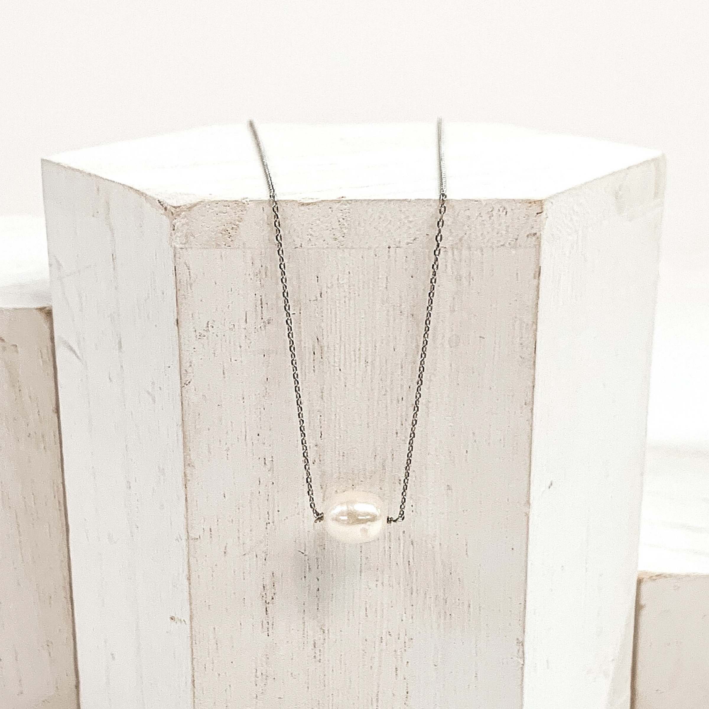 Simple, silver chain necklace with a single white pearl bead. Pictured laying on a white block on a white background. 