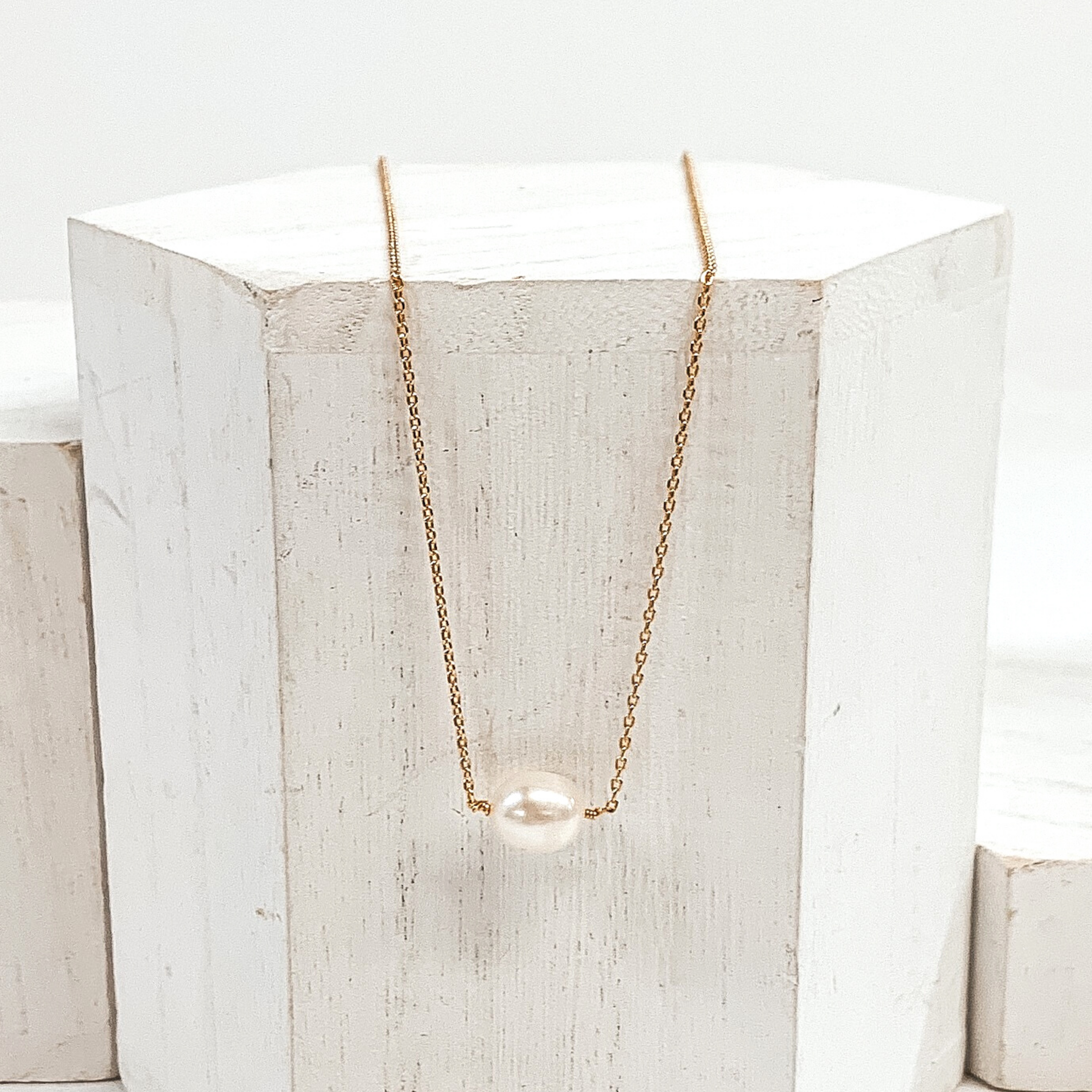 Simple, gold chain necklace with a single white pearl bead. Pictured laying on a white block on a white background.