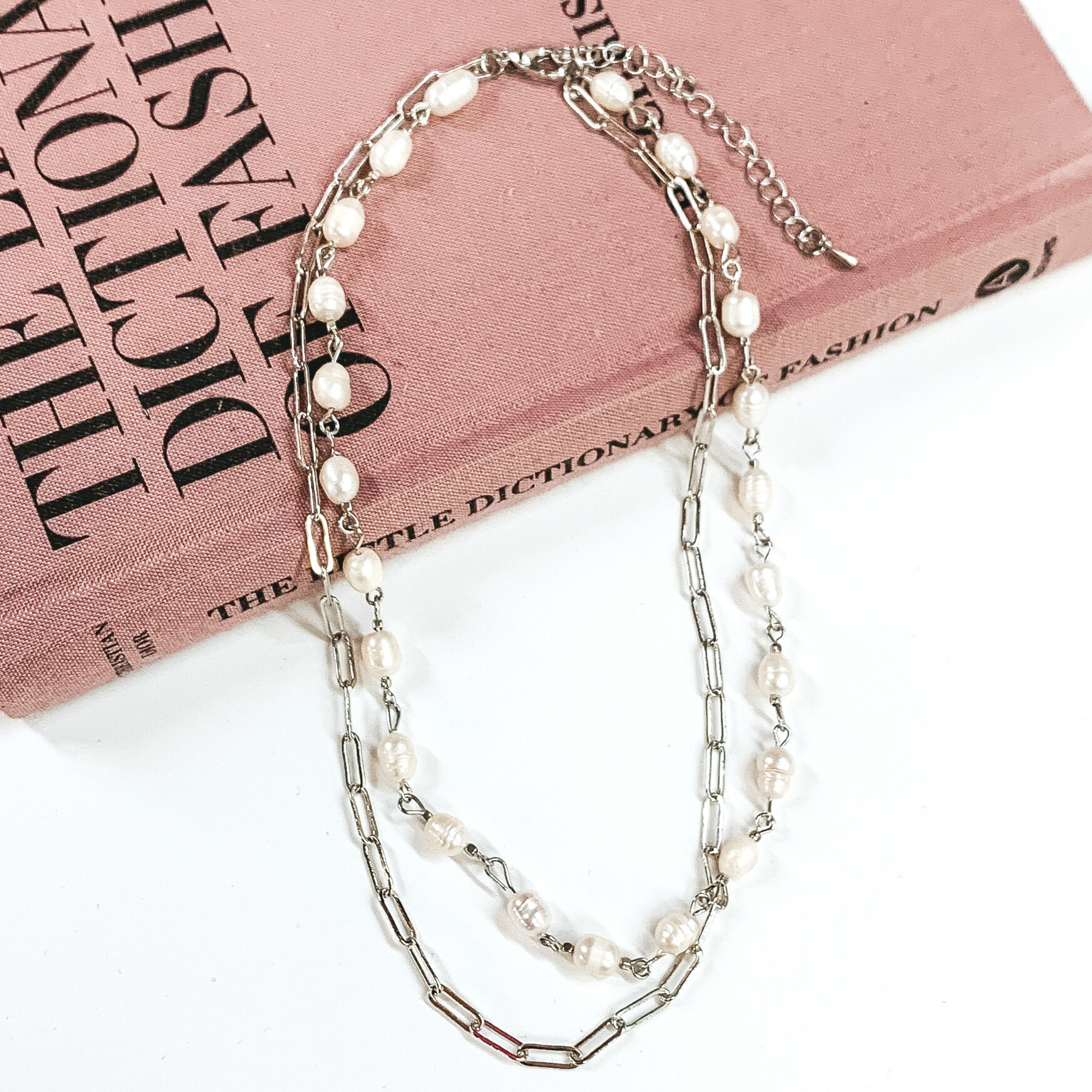 Silver, double layered necklace. One strand is a silver paperclip chain and the second strand is a white pearl beaded linked chain. This necklace is pictured laying partially on a mauve colored book on a white background. 