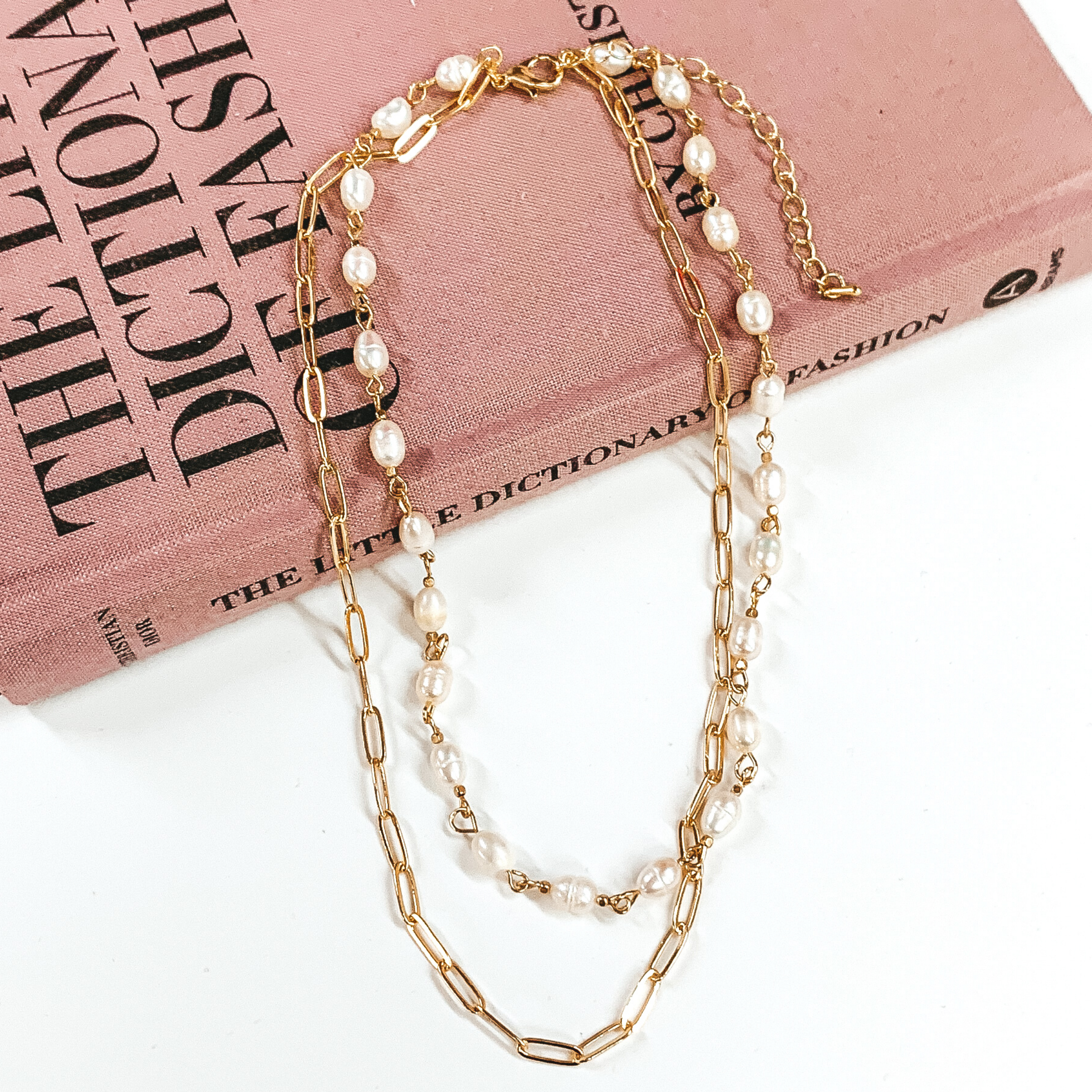 Gold, double layered necklace. One strand is a gold paperclip chain and the second strand is a white pearl beaded linked chain. This necklace is pictured laying partially on a mauve colored book on a white background. 