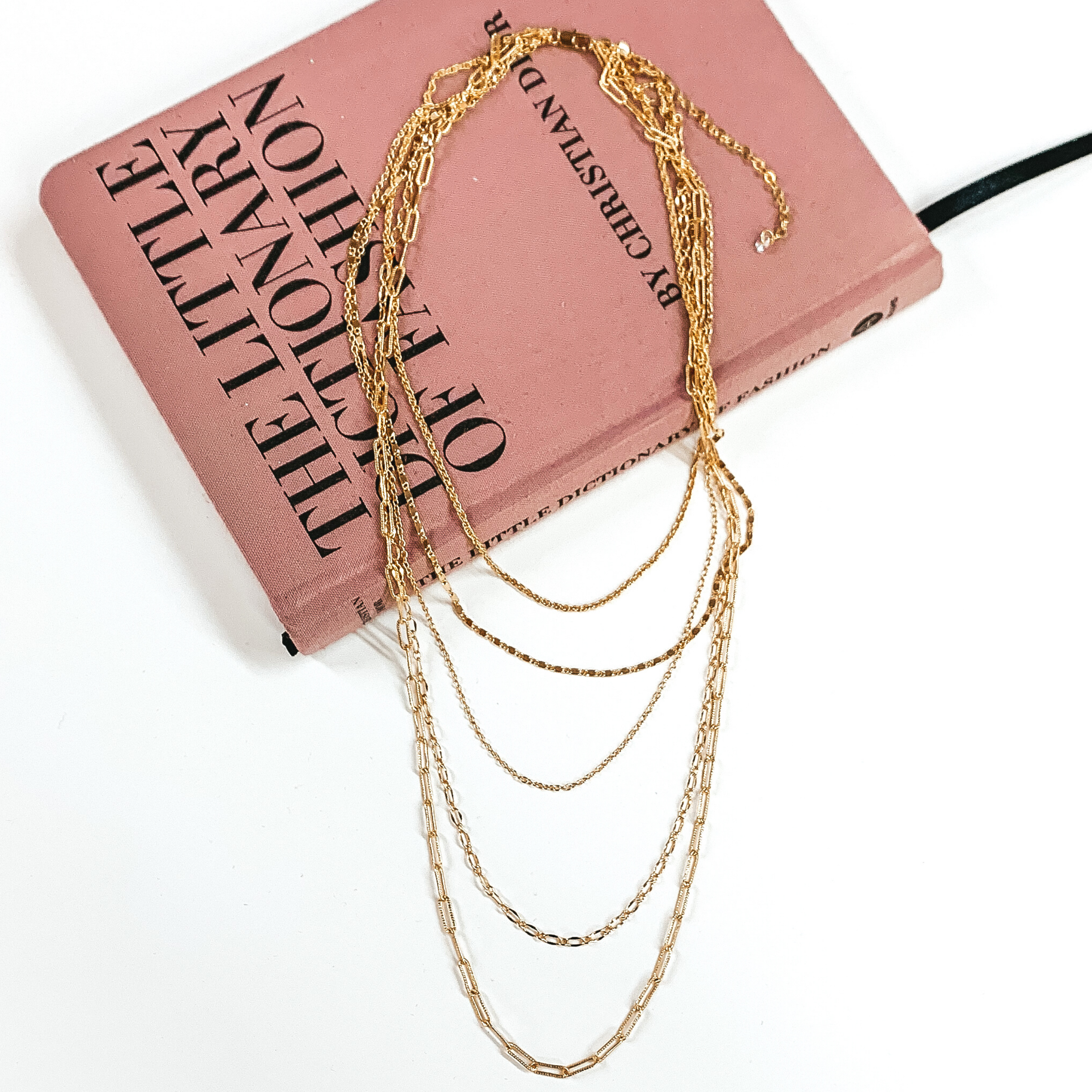 5 layered gold multi chained necklace. This necklace is pictured laying partially on a mauve colored book on a white background. 