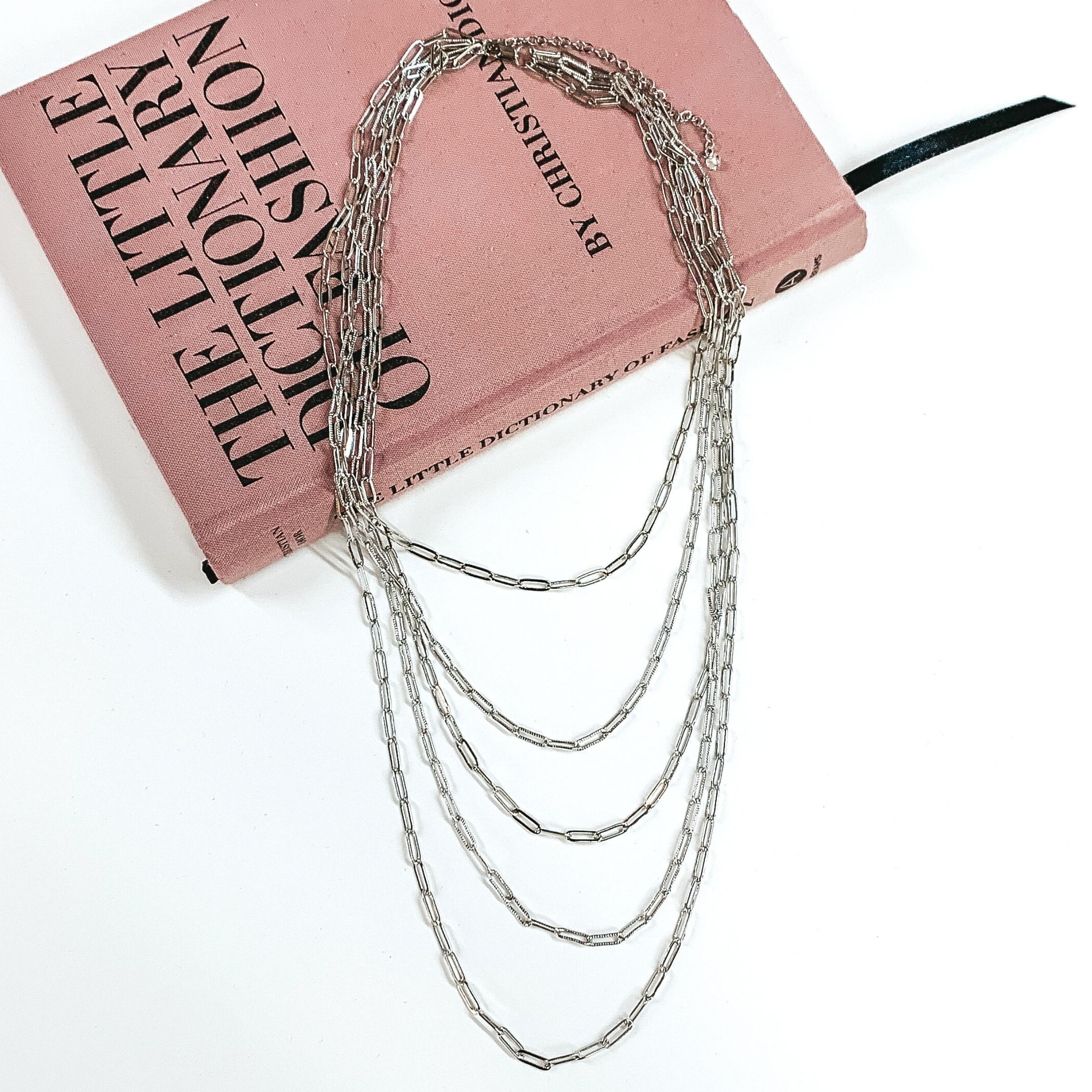 5 layered silver paperclip chained necklace. This necklace is pictured laying partially on a mauve colored book on a white background. 