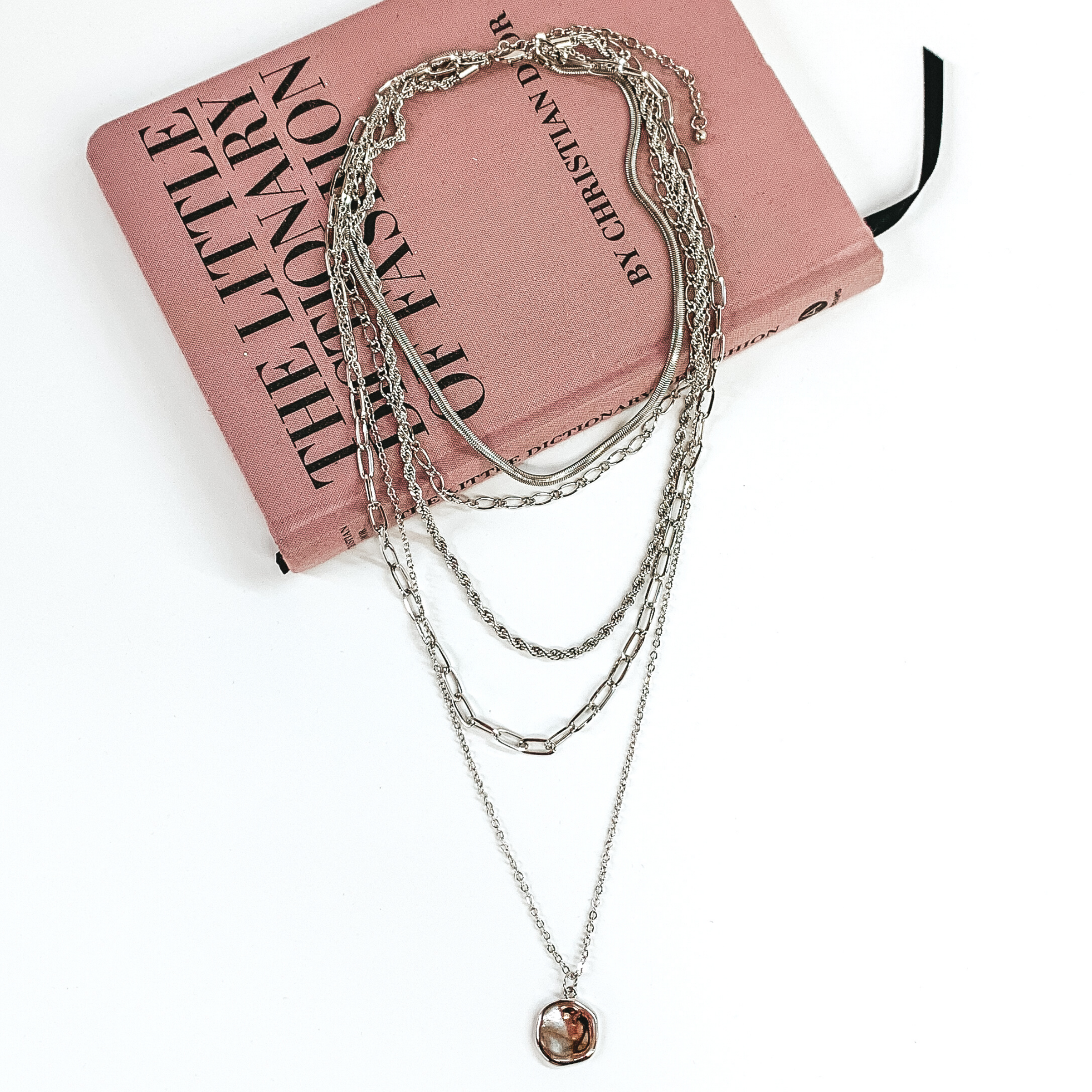 Silver multi chained necklace. This necklace has different types of chain for each strand and the longest strand has a circle pendant. This necklace is pictured laying partially on a mauve colored book on a white background. 
