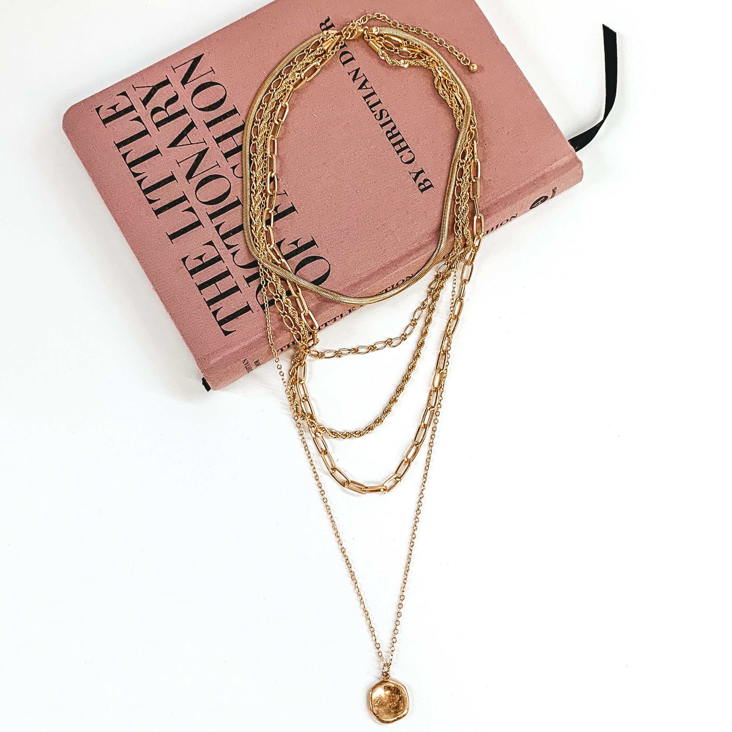 Gold multi chained necklace. This necklace has different types of chain for each strand and the longest strand has a circle pendant. This necklace is pictured laying partially on a mauve colored book on a white background. 