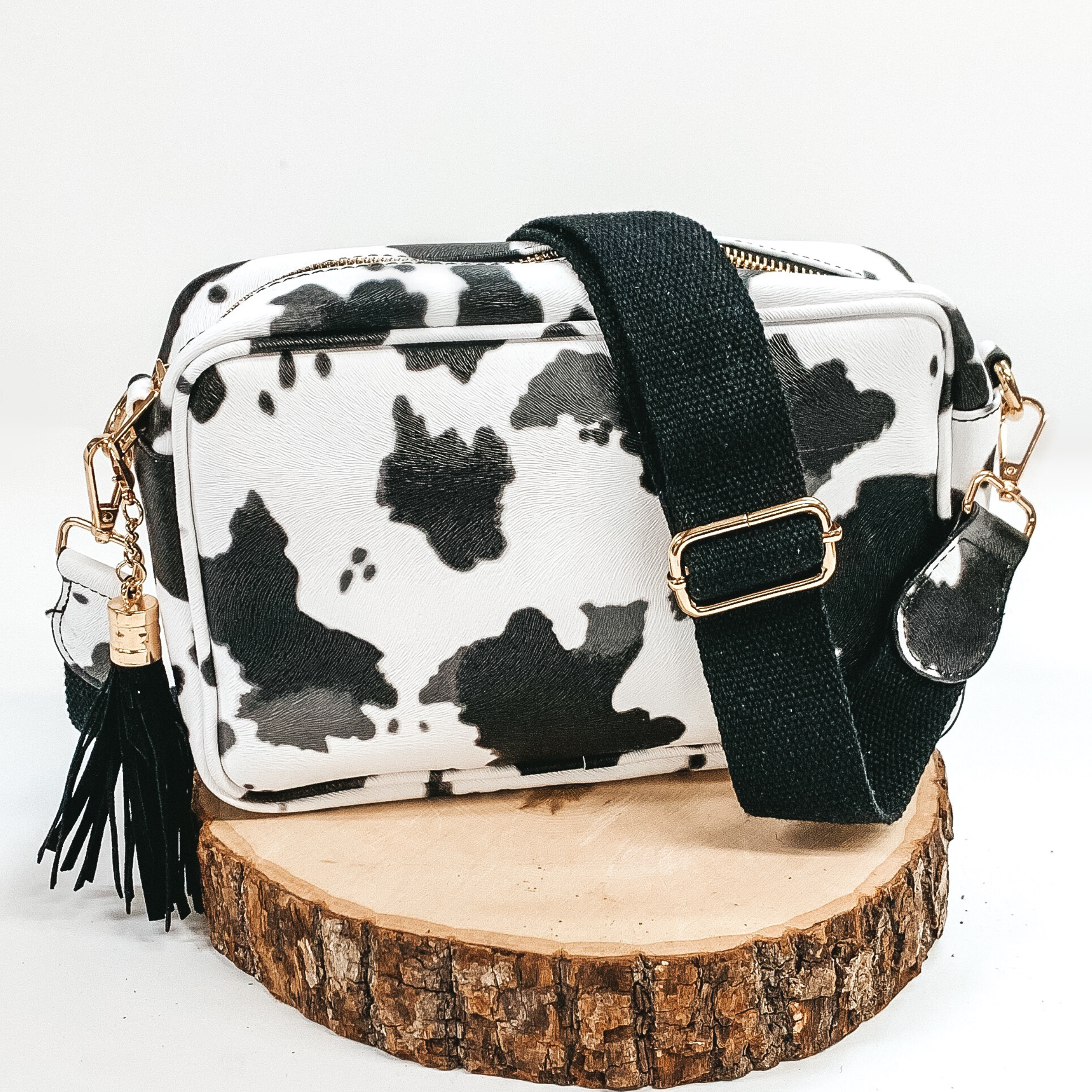 White and black cow print rectangle purse with a black adjustable strap and black decorative tassel. This purse also includes gold accents. This purse is pictured sitting on a piece of wood on a white background.