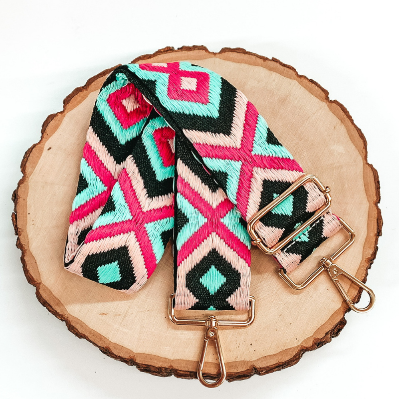 Black canvas purse strap with light pink, hot pink, and aqua embroidered design. This purse strap includes gold accents. This purse strap is pictured on a piece of wood on a white background. 