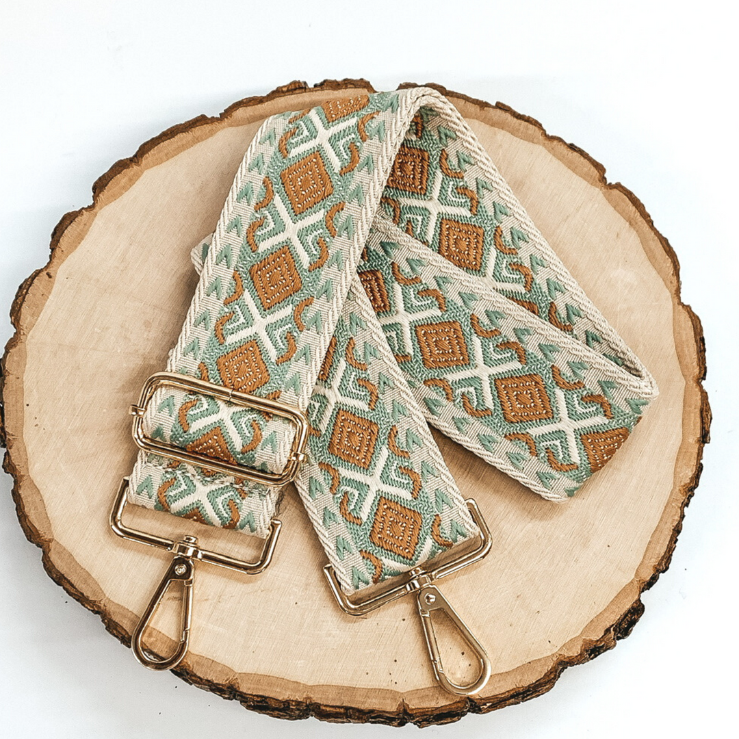 Beige canvas purse strap with tan and olive embroidered design. This purse strap includes gold accents. This purse strap is pictured on a piece of wood on a white background. 