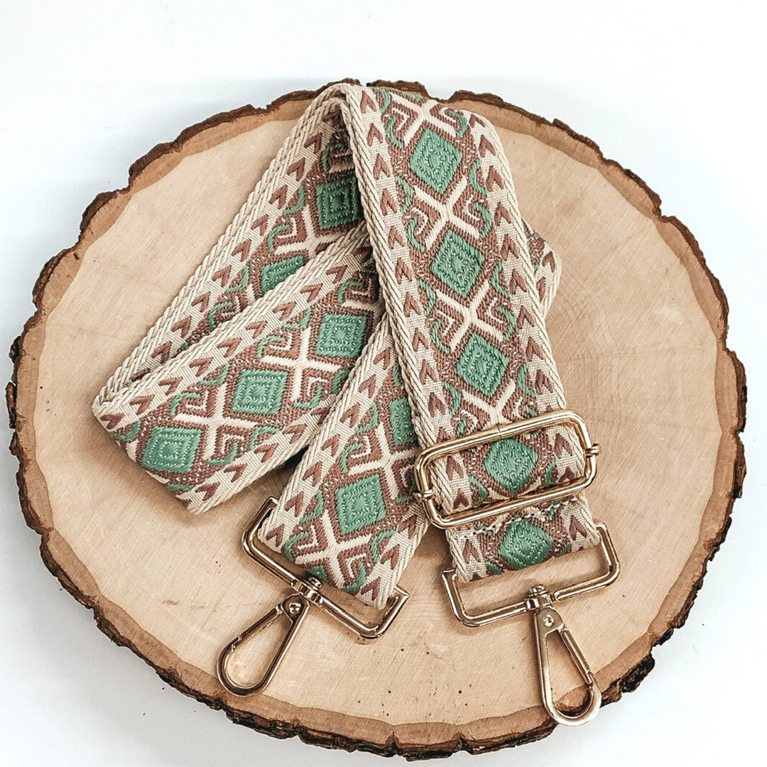 Beige canvas purse strap with mauve and olive embroidered design. This purse strap includes gold accents. This purse strap is pictured on a piece of wood on a white background. 