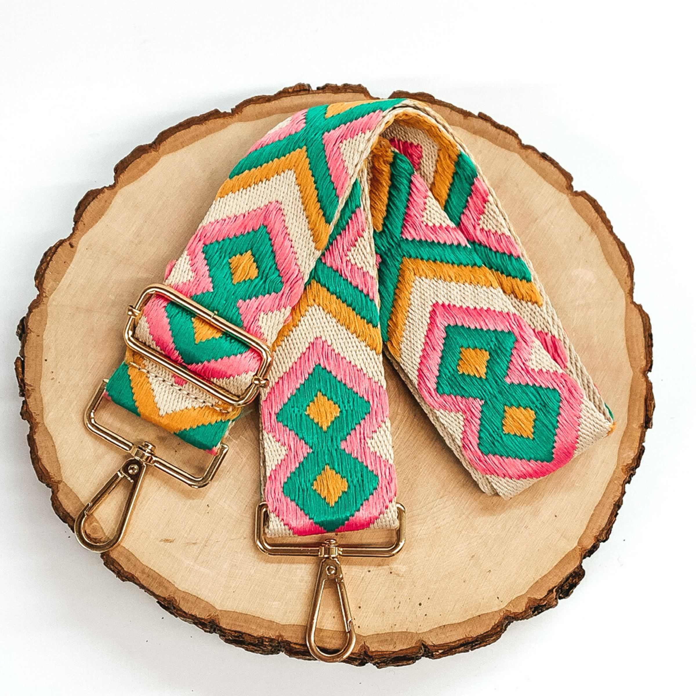 Beige canvas purse strap with yellow, pink, and teal embroidered design. This purse strap includes gold accents. This purse strap is pictured on a piece of wood on a white background. 
