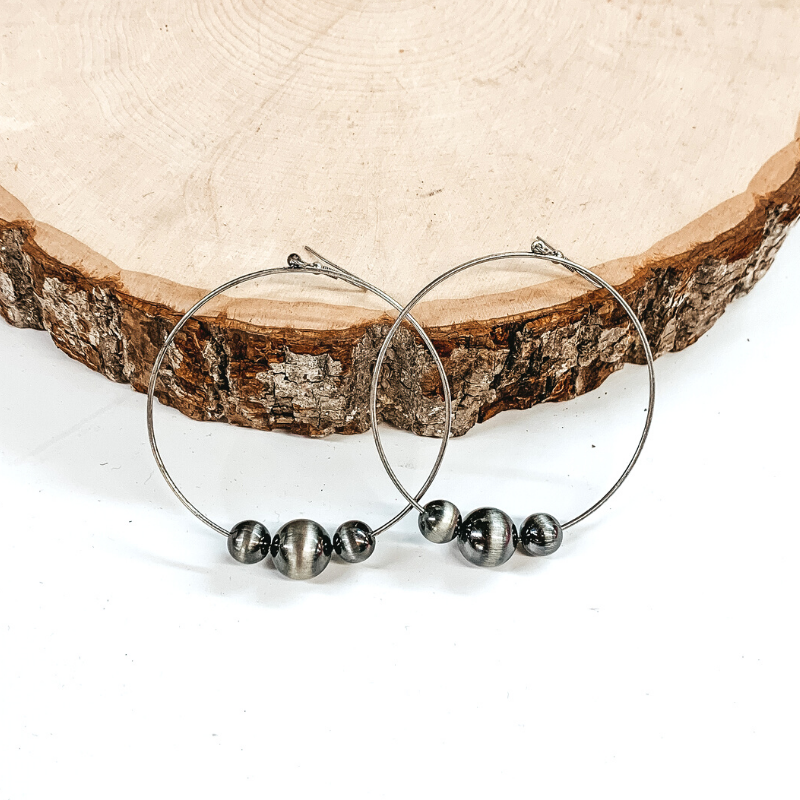 Silver open circle drop earrings with silver beads in varying sizes at the bottom of the earrings. These earrings are pictured laying against a piece of wood. 